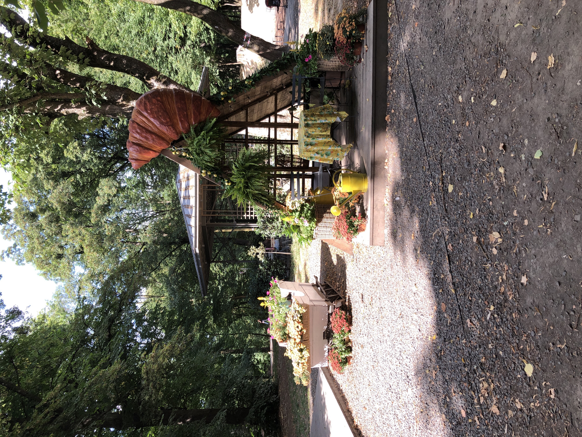 The image depicts an outdoor cafe setting in Lviv, Ukraine, on a sunny day. In the foreground, a gravel pathway is visible, with scattered leaves and shadows cast by trees. To the left, a light-colored, decorative carriage filled with an abundance of yellow and pink flowers sits on the gravel. Further to the right, a wooden deck platform hosts a small cafe setup. A triangular wooden structure with a metal roof serves as a canopy, adorned with a large, reddish-brown croissant sculpture and hanging greenery. Underneath the canopy, a round table is covered with a yellow tablecloth patterned with floral motifs, and two dark chairs are placed around it. A bright yellow watering can sits on the deck near the table, and various potted plants, including chrysanthemums in shades of orange and red, are arranged around the deck and the carriage. In the background, mature trees with lush green foliage line the area, suggesting a park-like environment. The overall scene conveys a whimsical and inviting atmosphere.