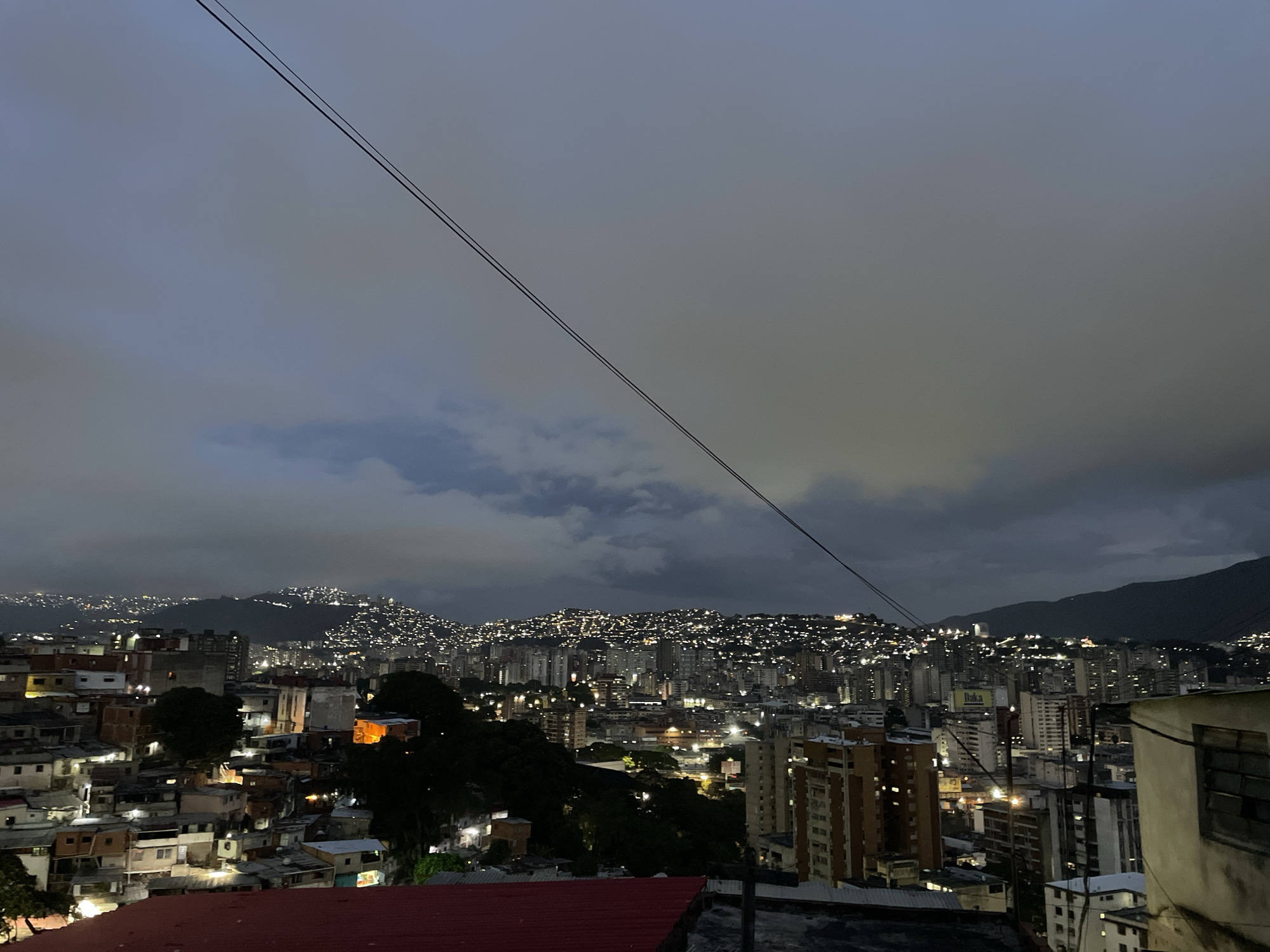 The image shows an elevated, nighttime view of Caracas, Venezuela. The foreground features densely packed, low-rise buildings, transitioning into a midground with taller structures characteristic of a city center. Numerous lights, likely from residences and street illumination, speckle the landscape. In the background, the landscape rises into hills, also dotted with lights, and a dark mountain range is silhouetted against the sky. A pair of utility lines cut diagonally across the upper portion of the frame. The sky is overcast, creating a soft, diffused light across the scene.