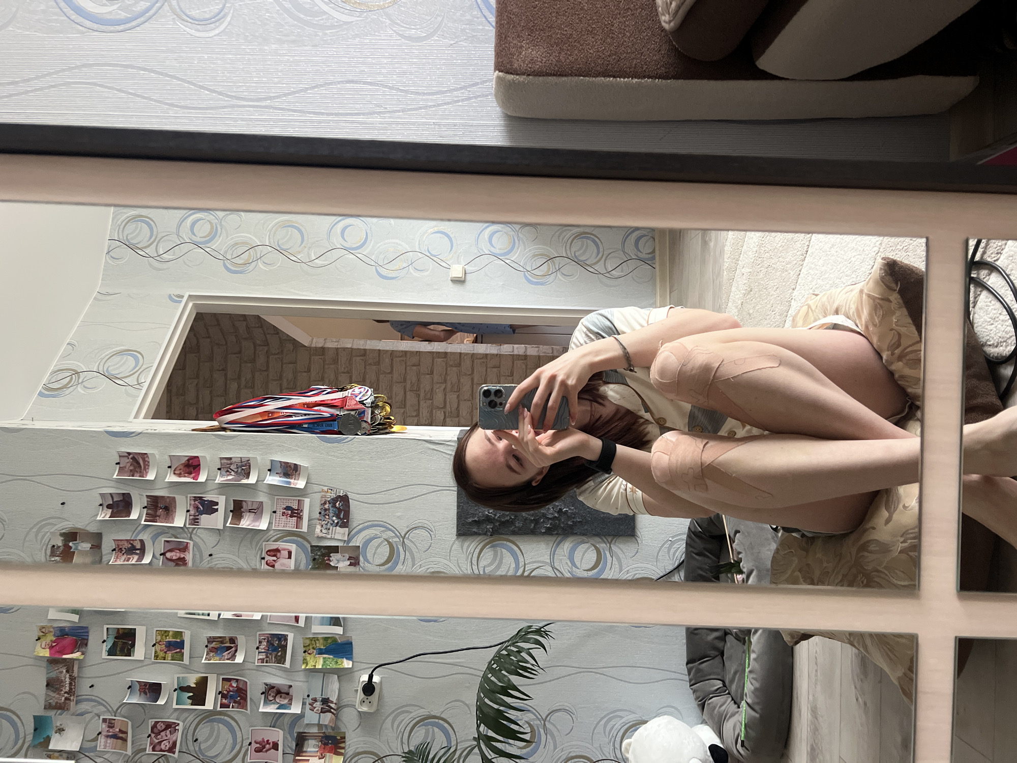 A young female is captured in a mirror selfie within what appears to be a residential interior in Chaikovsky, Russia. The image is taken through a large, possibly segmented mirror, reflecting the subject and parts of the room. The female, with long dark brown hair, holds a smartphone (likely an iPhone) to take the picture, with the device partially obscuring her face. She is dressed in light-colored clothing and is seated or reclining on a bed or sofa, accompanied by a patterned pillow.
A notable detail is the presence of several strips of medical tape (such as kinesiology tape) applied to both of her legs.
The room's decor includes light-colored wallpaper featuring a wavy, circular pattern in shades of blue, grey, and brown. Adorning the wall are multiple small, printed photographs arranged in rows, affixed with pins. A collection of medals, some with ribbons, hangs to the side, with the text "BIG KICK" faintly visible on one. Visible through a doorway in the background is another room with a brick-patterned wall. Other objects include a houseplant, a power outlet with a cable plugged in, a white light switch, and a white furry object (possibly a stuffed animal or blanket) on the floor/bed. The setting suggests a casual, personal space, likely a bedroom or living area.
