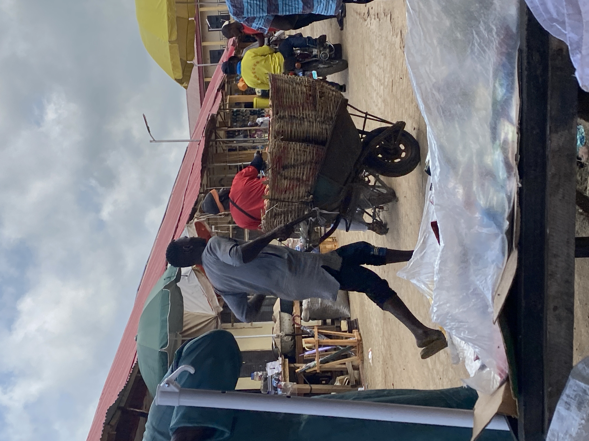 The image depicts a street scene in Kafanchan B, Nigeria. A man in a light gray shirt and dark shorts is pulling a heavily loaded wheelbarrow filled with what appears to be woven baskets. The setting is a marketplace; there are buildings with red roofing on the left, and several people are milling about, including individuals wearing yellow and red shirts. Another person is sitting on a motorcycle in the background. The area seems to be a busy marketplace with various stalls and objects visible, including umbrellas and assorted merchandise. The weather seems partly cloudy.