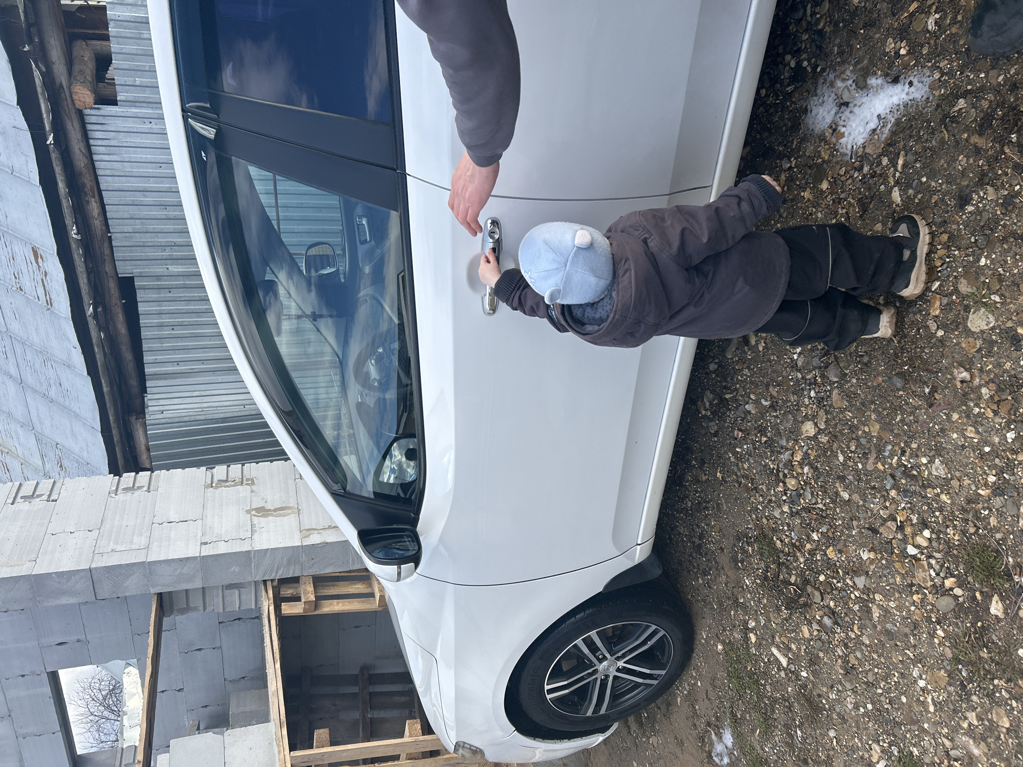The image, taken in Chaikovsky, Russia, depicts an outdoor scene featuring an adult and a small child interacting with a white car during daylight hours. A child, dressed in a dark puffer jacket, black trousers, and a light blue hat, is shown reaching for or holding the car's silver door handle. An adult's arm and hand are visible above the child's, positioned near the car door, possibly assisting or guiding the child.
The car is a white sedan or hatchback, parked on unpaved ground composed of dirt and gravel, with some patches of white substance, likely snow or foam, suggesting cool weather. In the background, there is a building under construction, featuring light-colored concrete blocks, wooden beams, and corrugated metal sheets. The car has multi-spoke alloy wheels. The sky appears bright and clear. There is no visible text in the image.