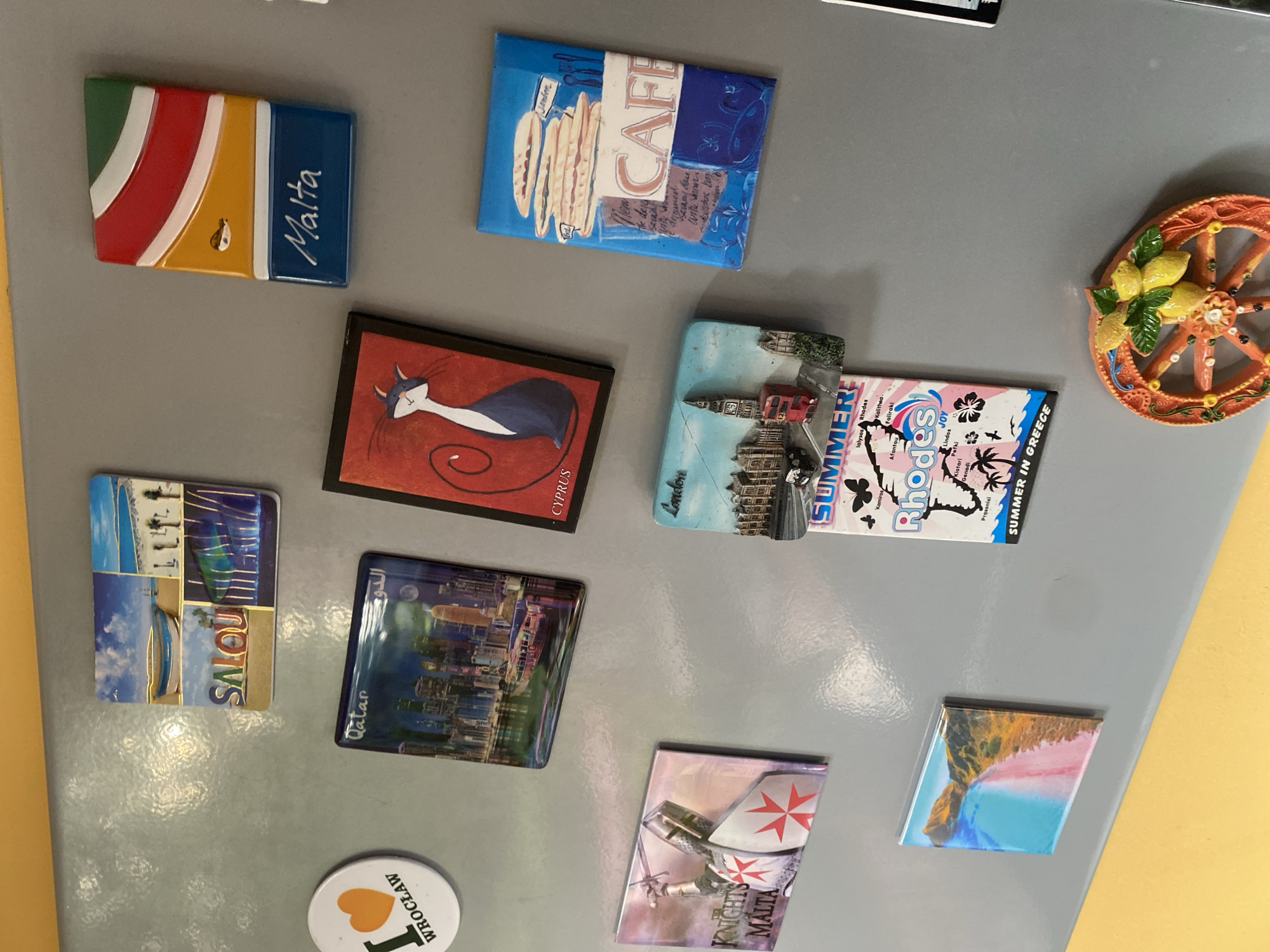 The image displays a collection of souvenirs, likely magnets, adhered to a refrigerator door. The souvenirs represent various locations. From top left, moving clockwise, are magnets from Salou, Malta, a magnet with "CAFE" written on it, a magnet featuring a stylized cat and the word "CYPRUS," a "SUMMER IN GREECE" magnet with "Rhodes" prominently featured, a magnet depicting a beach scene, a magnet of a knight with a shield, and a magnet from Qatar. Also visible is a circular white magnet with a red heart and the text "I ♡ WROCŁAW." The background is a grey, metallic surface, characteristic of a refrigerator door, with a yellow surface visible at the bottom. There are no people or animals present in the image. The lighting appears to be from overhead, casting some subtle reflections on the glossy surfaces of the magnets.