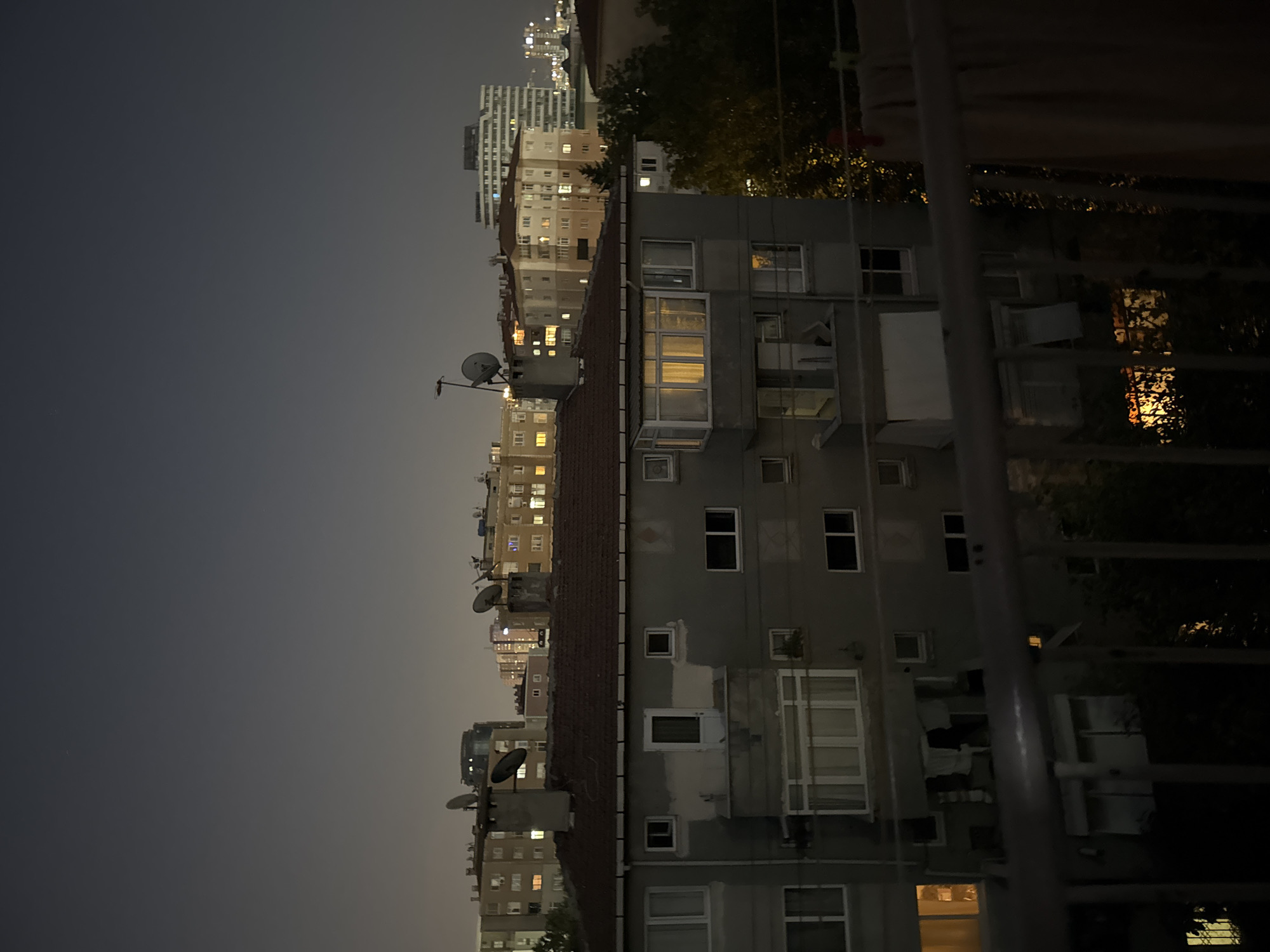 The image depicts a nighttime cityscape of Gungoren, Turkey, characterized by residential buildings and a dusky sky. In the foreground, a metal railing is visible, partially obscuring the view of a grey apartment building directly behind it. This building has multiple windows, some illuminated by warm interior lights, and a small balcony with hanging laundry. Above this building, other apartment complexes with lit windows are seen, along with satellite dishes mounted on rooftops. Further in the distance, a modern, white high-rise building stands out against the darker sky, along with other structures that appear to be in various stages of construction or illumination. The overall atmosphere is urban and residential, with the night setting casting a dim, ambient light.