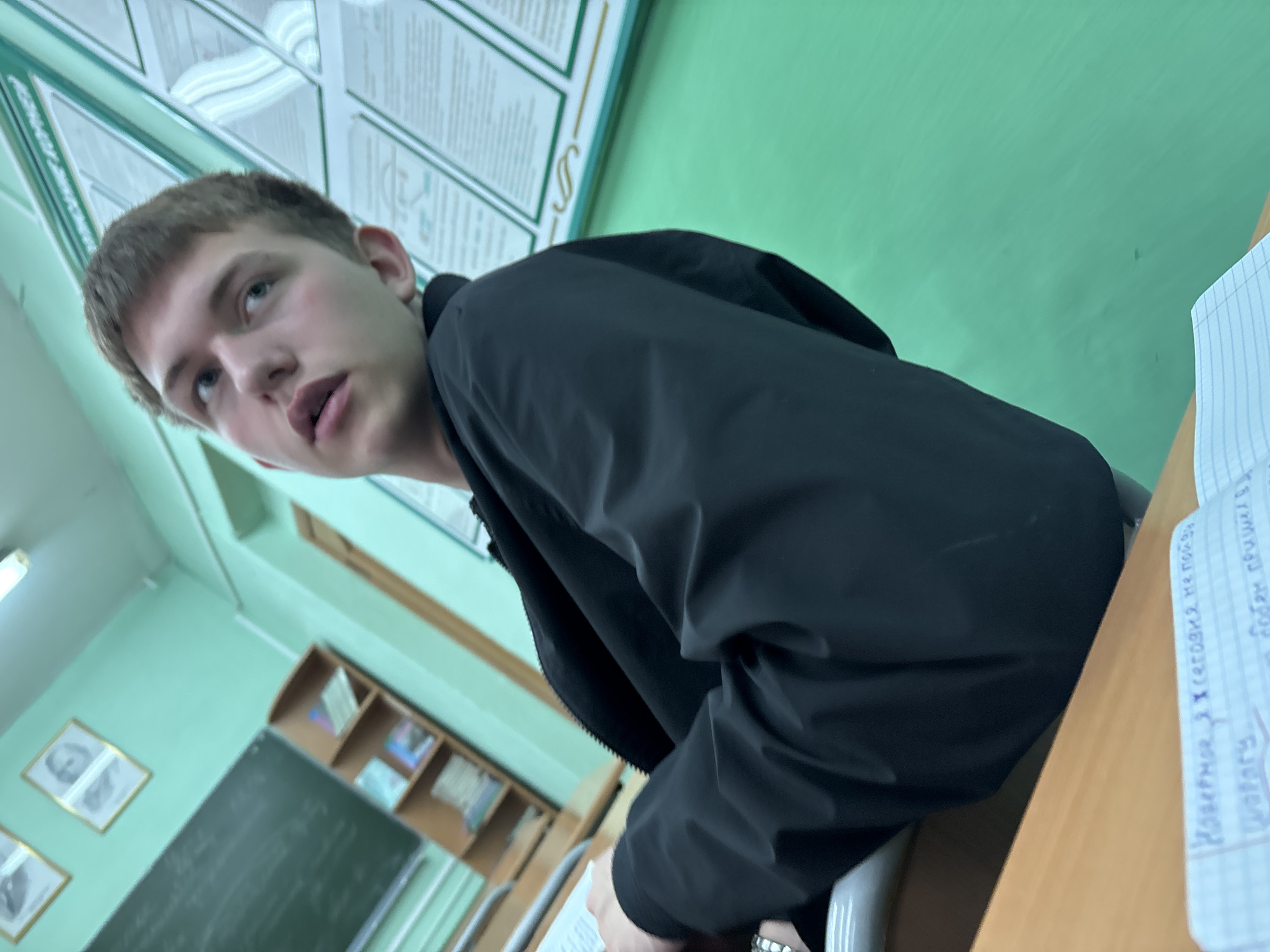 An indoor, tilted medium shot captures a young male in what appears to be a classroom setting. The subject, a person with short brown hair and light eyes, is positioned off-center, looking slightly upwards and to his left with a thoughtful or curious expression, his mouth slightly ajar. He is wearing a dark, possibly black, jacket.
He is seated at a wooden desk, and his hands are near a notebook with ruled paper, suggesting he might be studying or taking notes. The background features light green walls. On the right side of the desk, a notebook is visible with handwritten text in Russian. Partially decipherable phrases include: "Наверное X (сегодня не пойдешь)" (Probably X (today you won't go)), "добро пришёл в школу" (good came to school), and "дорогу." (road/way).
The classroom environment is further indicated by charts or posters with text on the wall behind the subject, a chalkboard, and shelves filled with books or other items in the blurred background. Framed pictures are also visible on a lower section of the wall. The lighting appears to be typical for an indoor setting during the day. The image is significantly rotated, giving a skewed perspective of the scene.