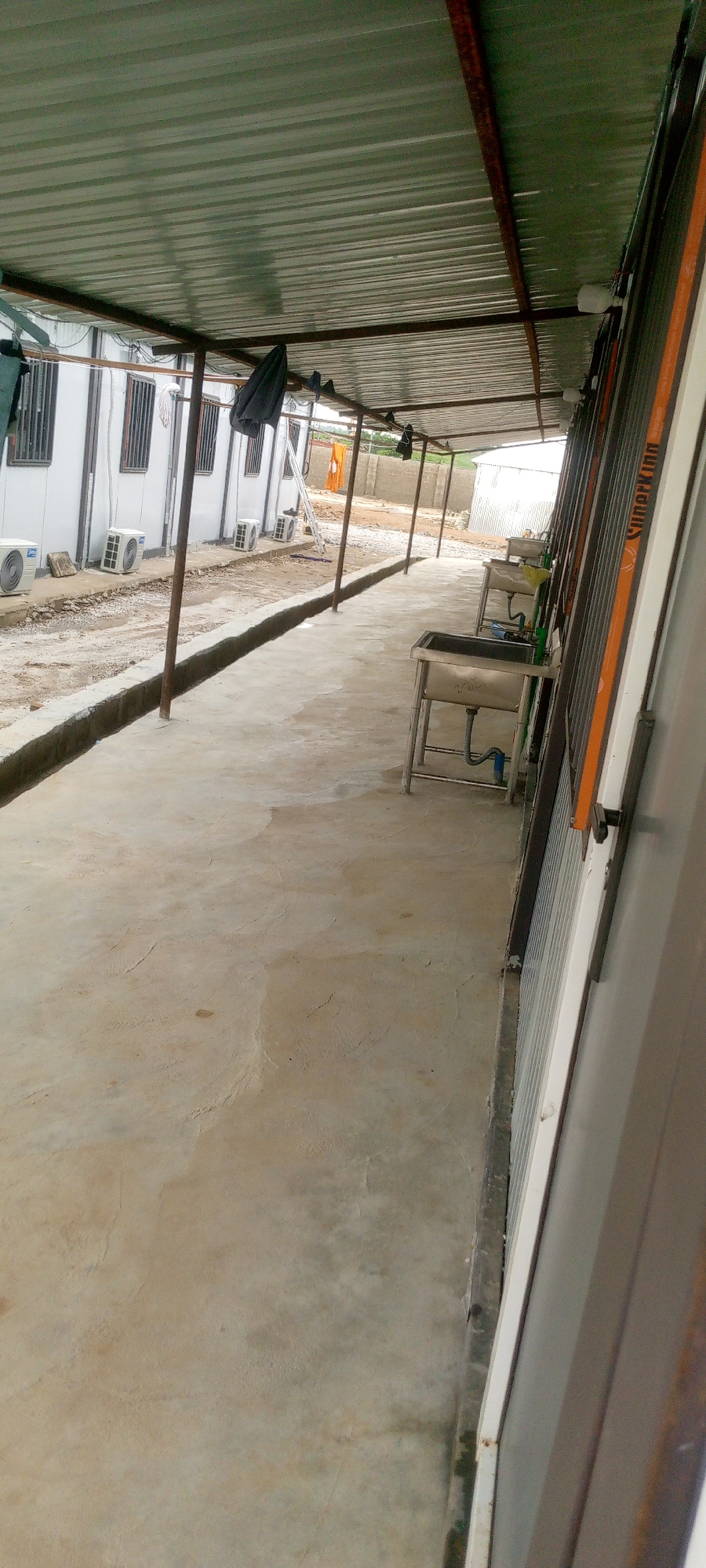 The image depicts an outdoor area adjacent to a row of modular buildings, likely in a construction or temporary settlement context. The scene is located in Indo, Nigeria. The foreground features a concrete pathway with a shallow, concrete-lined trench running along its edge. To the left, a white modular building is visible, with several air conditioning units mounted on its exterior. The building has windows with dark grills. A corrugated metal roof, supported by rusty metal poles, extends over the pathway, providing shade. Several dark-colored items, possibly clothing, are hanging from the roof supports.
Further down the pathway on the right side, there are two stainless steel sinks with metal stands, plumbing, and faucets. These appear to be part of an outdoor wash station. The background shows more of the modular buildings, a fence made of corrugated metal or wire, and an open, somewhat dusty area. A ladder is leaning against one of the buildings. The overall impression is one of functional, perhaps temporary, outdoor infrastructure.