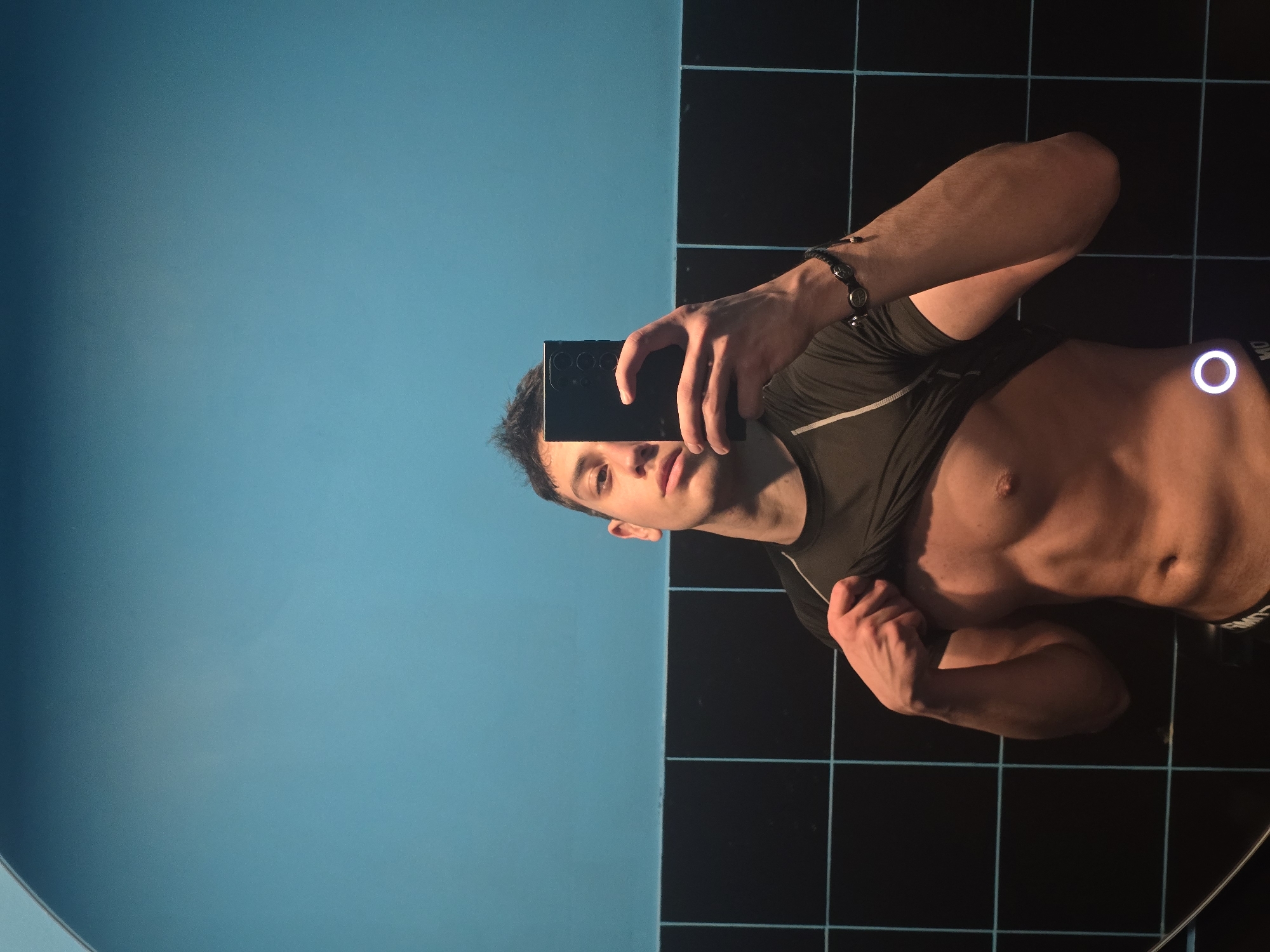 A man is taking a selfie in what appears to be a bathroom setting. He is holding a smartphone in his right hand, obscuring part of his face. He is wearing a dark-colored athletic shirt that is partially pulled up, revealing a portion of his torso. The background consists of a blue wall and a grid of dark tiles with light-colored grout lines. The location in the city of Yerevan, Armenia, is unknown.