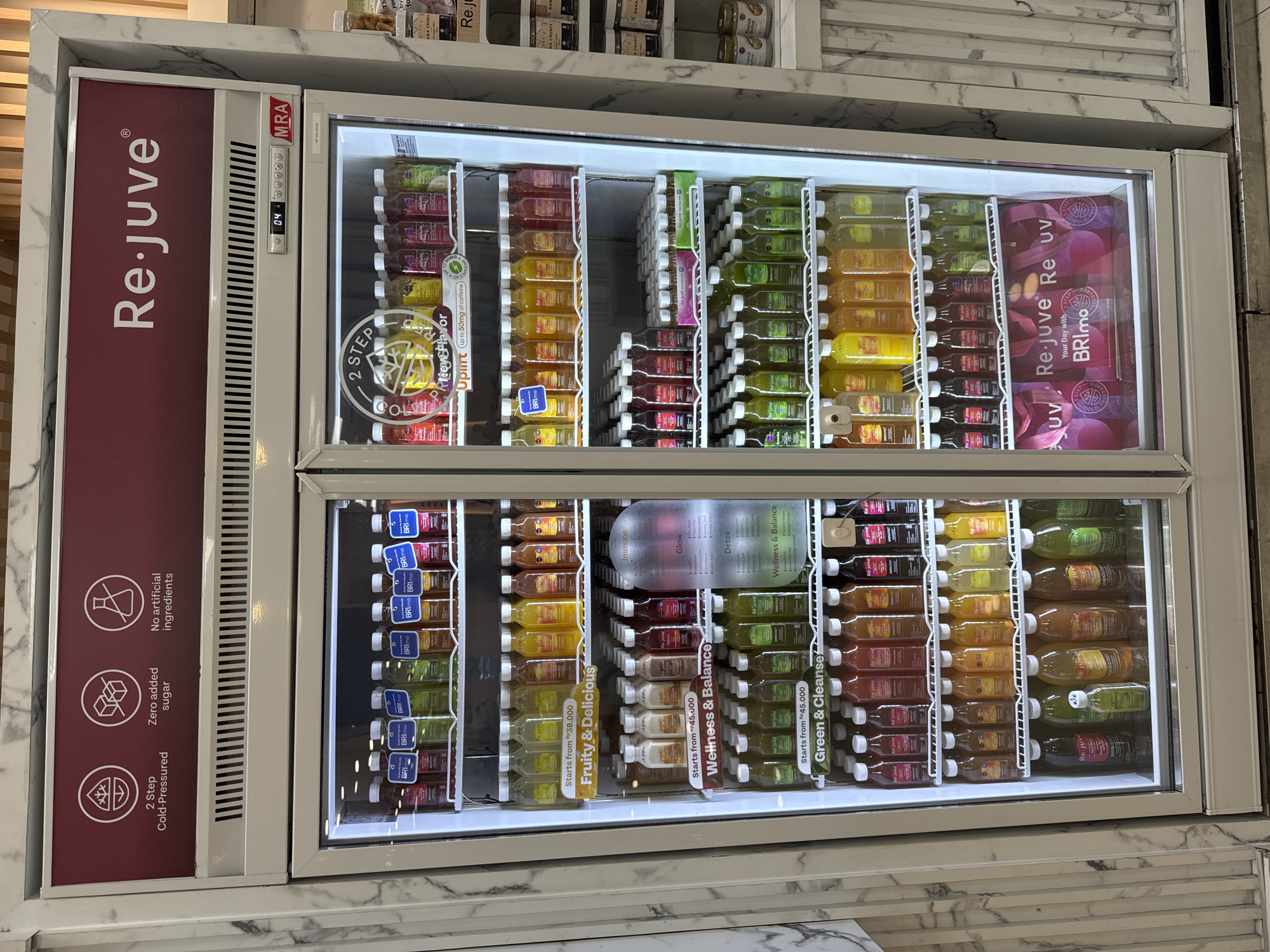 In Kuta, Indonesia, a full-length shot shows a refrigerated display case filled with bottled juices from the brand "Re.juve". The refrigerator is a white, double-door unit with a prominent "Re.juve" logo at the top, alongside icons indicating "2 Step Cold-Pressed", "Zero added sugar", and "No artificial ingredients". The digital temperature display shows "04".
The refrigerator is organized into multiple shelves containing numerous bottles of juice, color-coded by flavor or type. Labels such as "Fruity & Delicious", "Wellness & Balance", and "Green & Cleanse" are visible, along with price indicators starting from "Rp38,000" and "Rp45,000". Some shelves also have accompanying informational signs or descriptive text.
Below the main display, the bottom section of the refrigerator reveals branded "Re.juve" shopping bags in pink and purple hues, with text like "Your Day with BRImo". The overall environment appears to be an indoor retail space, possibly a health food store or a juice bar, with a white, marble-patterned wall visible on the left side of the image. No people are present in the frame.