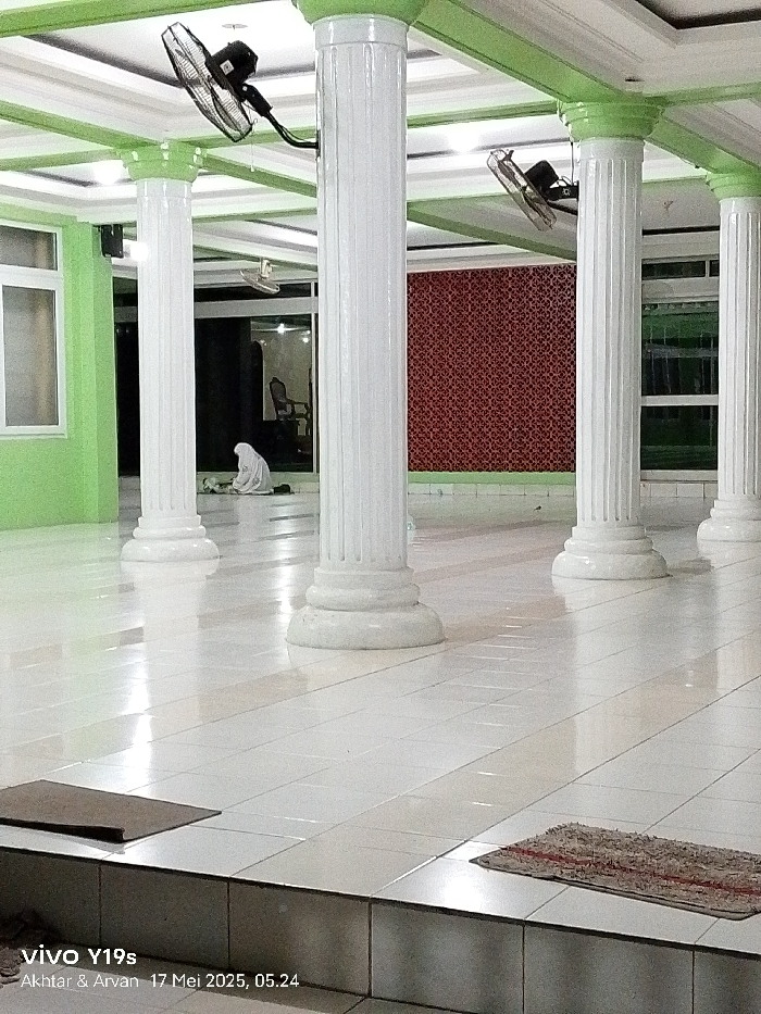 The image shows an interior space with white tiled floors and white pillars. There is one person in a white garment praying in the background, facing a wall with a red and white pattern. The ceiling has white and green trim. The image is taken in Gegesik, Indonesia, at the coordinates -6.536 latitude and 108.431 longitude. Two ceiling fans are visible in the image. The image was captured with a vivo Y19s phone. The time stamp on the image is 17 Mei 2023, 05:24.