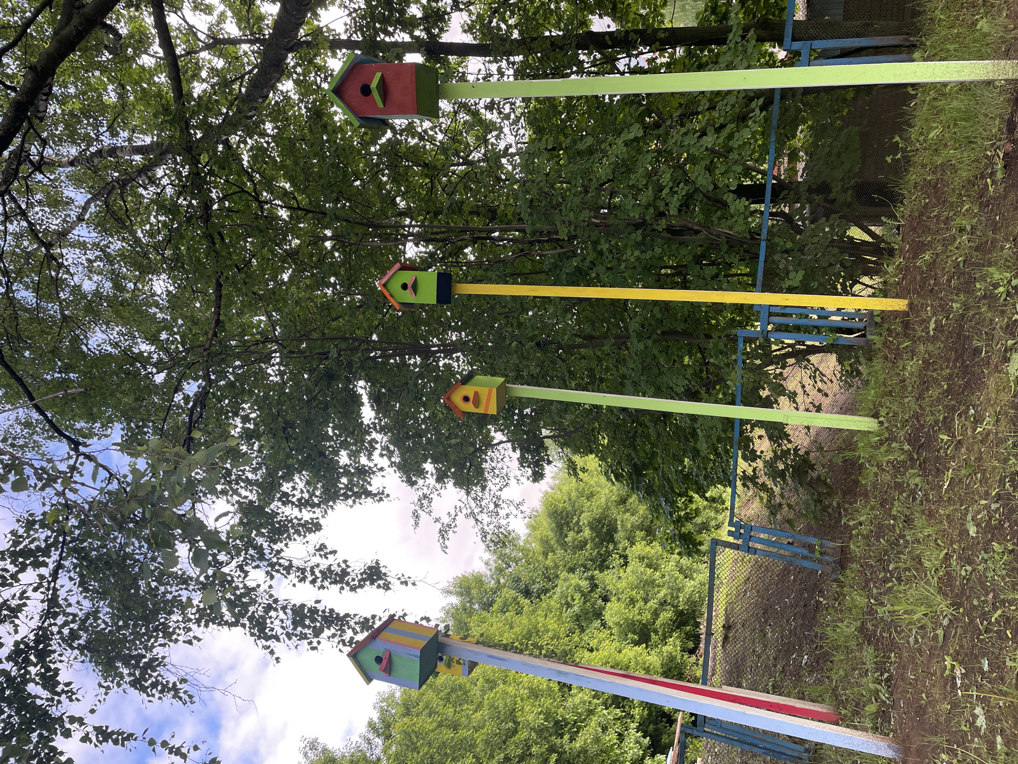 The scene depicts four brightly-colored birdhouses mounted on painted poles in a park-like setting in Izhevsk, Russia. The birdhouses are evenly spaced and appear to be part of a park installation or art display. The poles supporting the birdhouses are painted in different colors, including green, yellow, and a striped combination of white, red, and blue. The birdhouses are varied in design, each with a different color scheme and shape. The background consists of lush green trees, forming a dense canopy overhead, and a glimpse of the sky is visible through the foliage. A blue metal fence is partially visible in the foreground, near the base of the birdhouse poles. The ground is covered with grass and some bare patches of soil. The lighting suggests it is daytime, with filtered sunlight reaching the ground. There are no people visible in the frame.