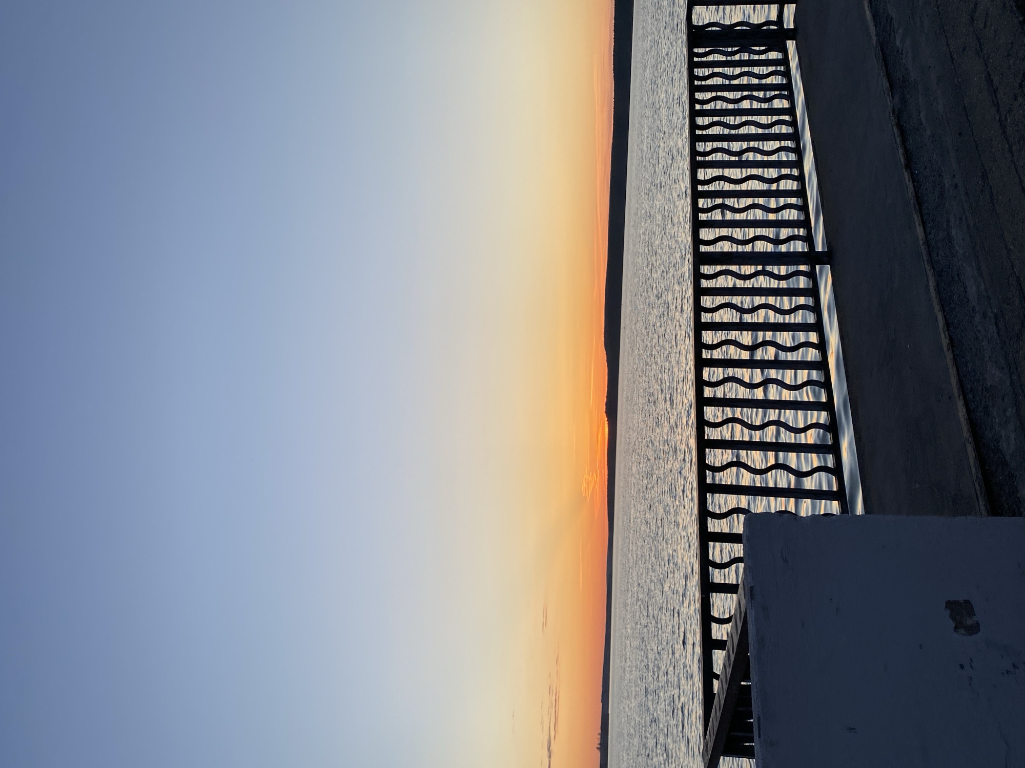 The image captures a view of a sunset over the Votkinsk Reservoir in Votkinsk, Russia. The sky transitions from blue to orange as the sun sets on the horizon. The reservoir appears rippled, reflecting the colors of the sky. On the right side, a metal staircase with a wavy pattern and concrete steps leads down to the water. A portion of a white, square structure is visible in the lower right corner.