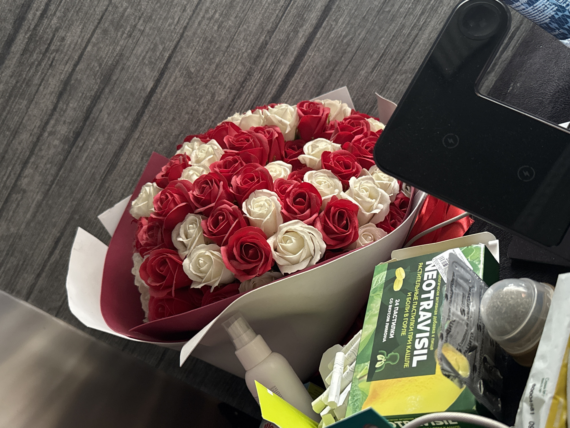 A close-up, angled shot shows a bouquet of red and white roses wrapped in red and white paper, placed on a textured, dark gray surface. To the left of the bouquet, a white spray bottle and a green and yellow box with Russian text and an image of a lemon are visible. Below these items, a black electronic device with two circular indentations and a circular lens is positioned in the foreground. The scene is lit from above, casting shadows on the surface and objects.