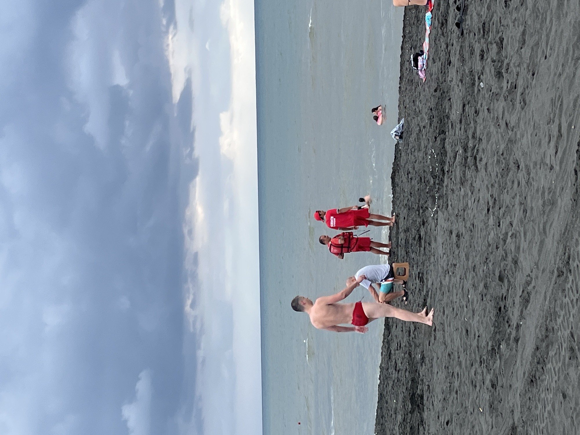 This image depicts a cloudy day on the black sand beach of Ureki, Georgia. In the foreground, a man wearing red swim trunks stands facing away from the camera, looking towards the sea. To his left, another man is seated on a small wooden stool, wearing a light blue t-shirt and shorts. In the middle ground, two individuals in red lifeguard uniforms are present, one wearing a life vest. Further out in the ocean, several people are visible swimming, including two individuals near a pink inflatable ring. The sea is calm, with gentle waves reaching the shore. The sky is overcast with dark, heavy clouds, suggesting a possibility of rain. Several towels and other beach items are scattered on the sand in the mid-ground and right side of the frame. The overall scene captures a typical day at a Georgian seaside resort.