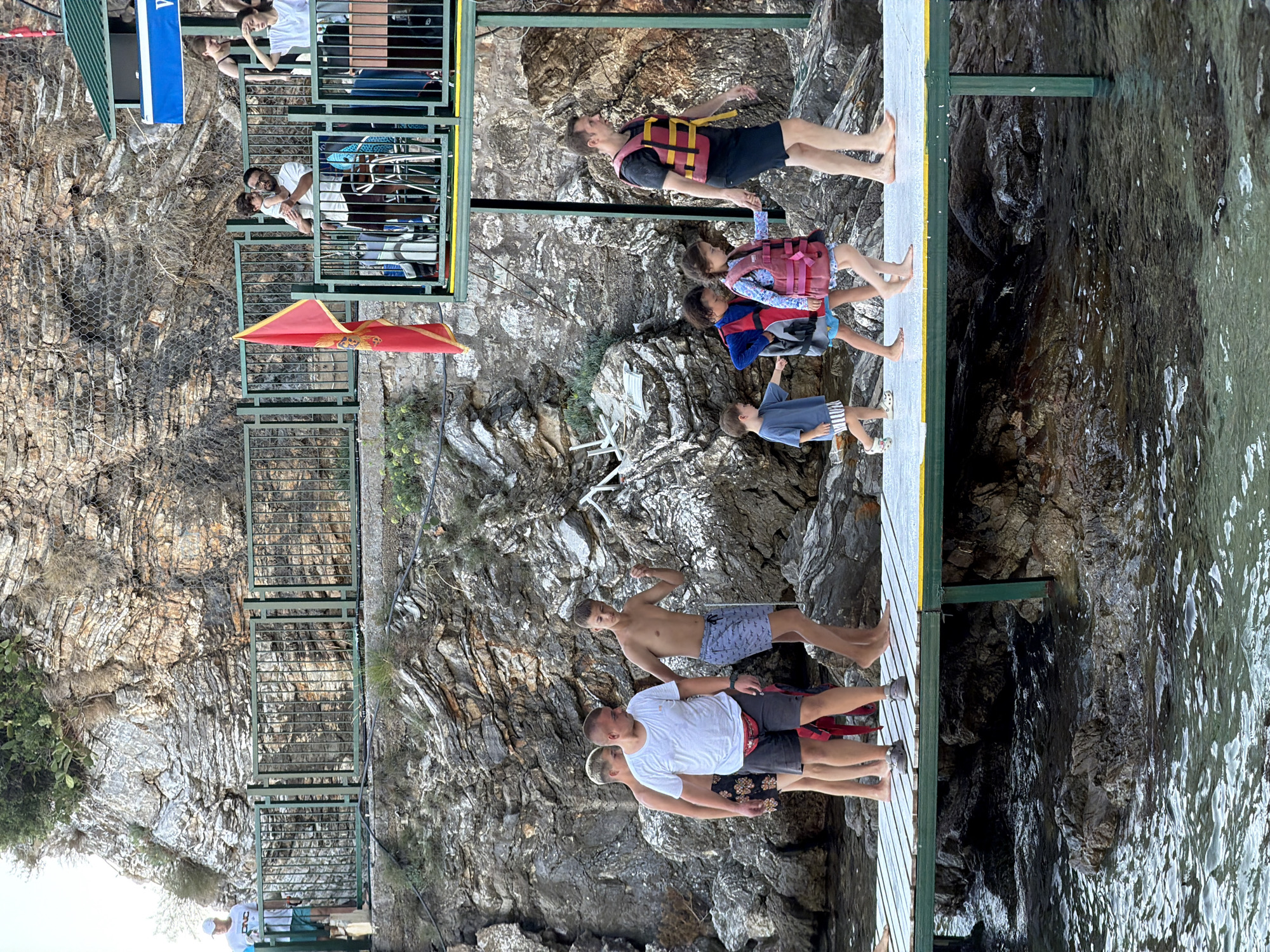 A group of people are walking on a wooden pier over the sea in Budva, Montenegro, with a rocky cliff face as the backdrop. On the pier, several men and boys are visible. Towards the left, three young men in swimwear are walking. One is wearing a white t-shirt and grey shorts with a red fanny pack. To his right, another young man is walking, and behind them, a third young man. Further along the pier, a man in a black t-shirt and dark shorts, wearing a yellow and red life vest, is walking, holding the hands of two young children. A young boy in a blue t-shirt and striped shorts is walking ahead of them. A young girl in a pink patterned swimsuit and a pink life vest is walking between the man and the boy. In the background, a red flag is attached to a pole. Above the pier, a higher platform with a railing is visible, where more people are standing and watching. The sea below the pier is clear and shows rocks. The environment is a coastal area with steep rock formations.