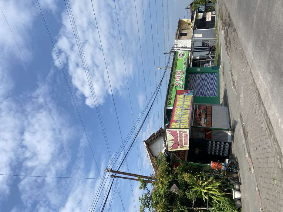 The image shows a street scene in Kecamatan Tanggulangin, Indonesia (latitude: -7.499, longitude: 112.7). There are several buildings along the street, including a small restaurant with a green sign that reads "RM. PUTRI WIKING MASAKAN PADANG". The restaurant has a white awning and a few tables and chairs outside. There is also a sign for "BAKSO & MIE AYAM PANGSIT" with a price of "7,000" displayed. In the foreground, there is a red bicycle leaning against the building. The street is paved with brick and is relatively empty of traffic. The sky is blue with some white clouds. There are multiple electrical lines crisscrossing the scene. The image was captured in the daytime.