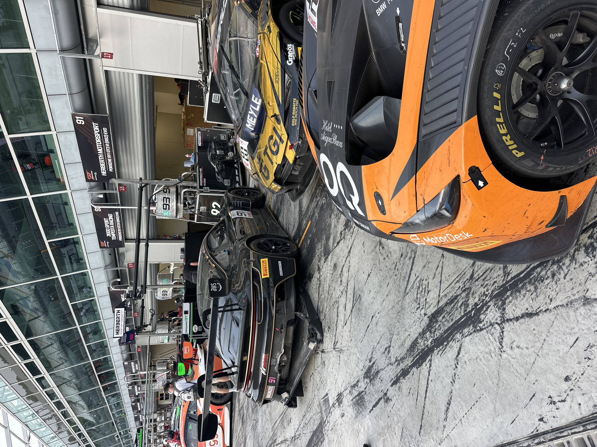 The image shows a pit lane at a racetrack, likely the Autodromo Nazionale Monza in Monza, Italy, given the presence of racing cars. Several racing cars are visible, including a black and gold car with "GTGI" and "MEYLE" logos, a black and orange BMW marked with "Hotel Aqua Bava," and a black car with the number 91. The garage bays behind the cars are marked with numbers such as 18, 19, 20, 21, and labels for "HERBERTH MOTORSPORT" and "ZIGGO SPORT TEMPESTA RACING." Signs indicate the types of cars being supported in these garages, such as "PORSCHE 911 GT3 R (992) EVO." Various racing support equipment is visible in the garages. The track surface appears worn and dirty. Some personnel are present in the garages, appearing to be working on the cars.