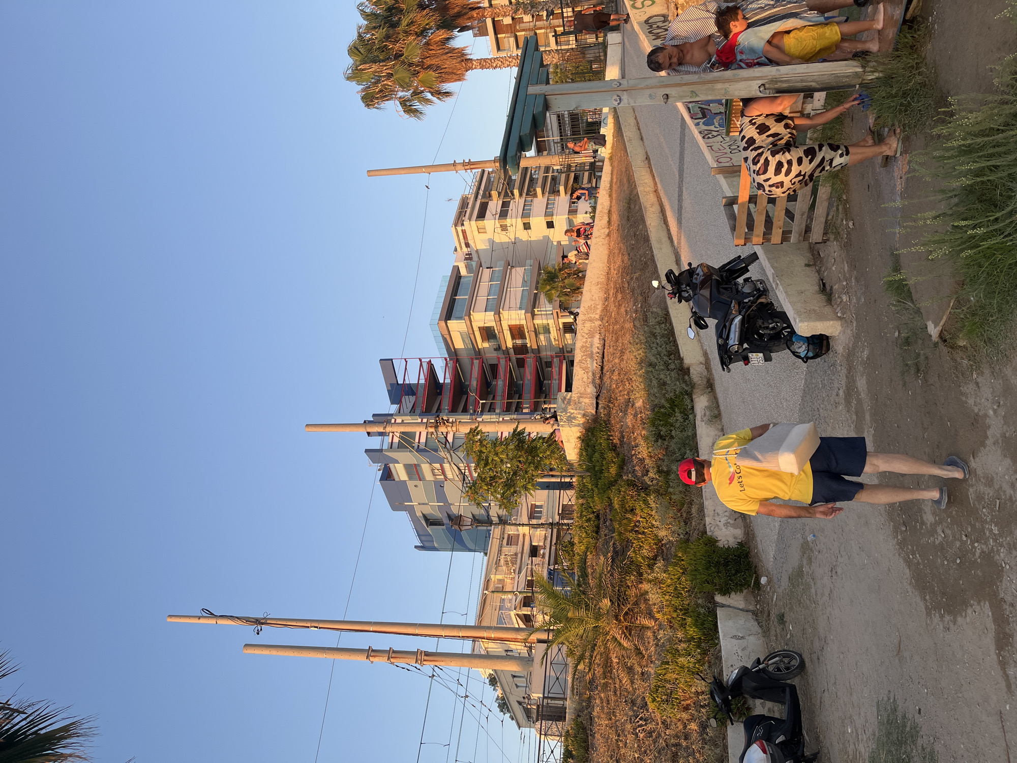 In the city of Paleo Faliro, Greece, during the golden hour, the scene captures a vibrant urban landscape transitioning into evening. The foreground is dominated by a dirt path with scattered debris, leading to a paved area where several people are engaged in various activities. A man in a yellow t-shirt and dark shorts walks away from the camera, carrying a large tote bag over his shoulder. To his left, a black scooter is parked. To the right, a group of individuals are gathered near an outdoor shower facility. A woman with a distinctive cow-print outfit is bending over, seemingly cleaning something, while a man in a striped shirt stands behind her. A child in yellow shorts is also present near the shower. The middle ground features a sloping pathway and a concrete retaining wall adorned with graffiti. In the background, a series of modern buildings with balconies and colorful architectural elements stand against a clear blue sky. Palm trees are visible, adding to the Mediterranean atmosphere. Overhead, utility poles and electrical wires indicate the presence of infrastructure. The warm light of the setting sun casts long shadows and illuminates the scene, creating a picturesque ambiance.