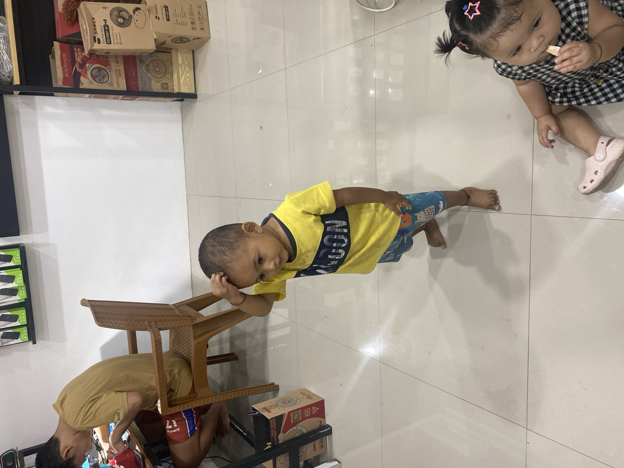 In a well-lit room, possibly a retail space in Ta', Indonesia, two young children are the central focus. A toddler, wearing a yellow t-shirt with a dark blue stripe and blue patterned shorts, stands looking down with his hand on his head, appearing contemplative or perhaps experiencing mild discomfort. To his right and slightly lower, a baby girl in a black and white checkered dress sits on the tiled floor, actively engaged with a small object she is holding in her hand and bringing to her mouth. The floor is made of light-colored ceramic tiles, reflecting the ambient light. In the background, to the left, a man is seated at a desk, seemingly working on a device, with a chair beside him. Shelves holding various boxes, some with fan imagery and branding such as "SANEX," are visible along the right wall. The environment suggests a commercial setting, perhaps an electronics store or a similar business.