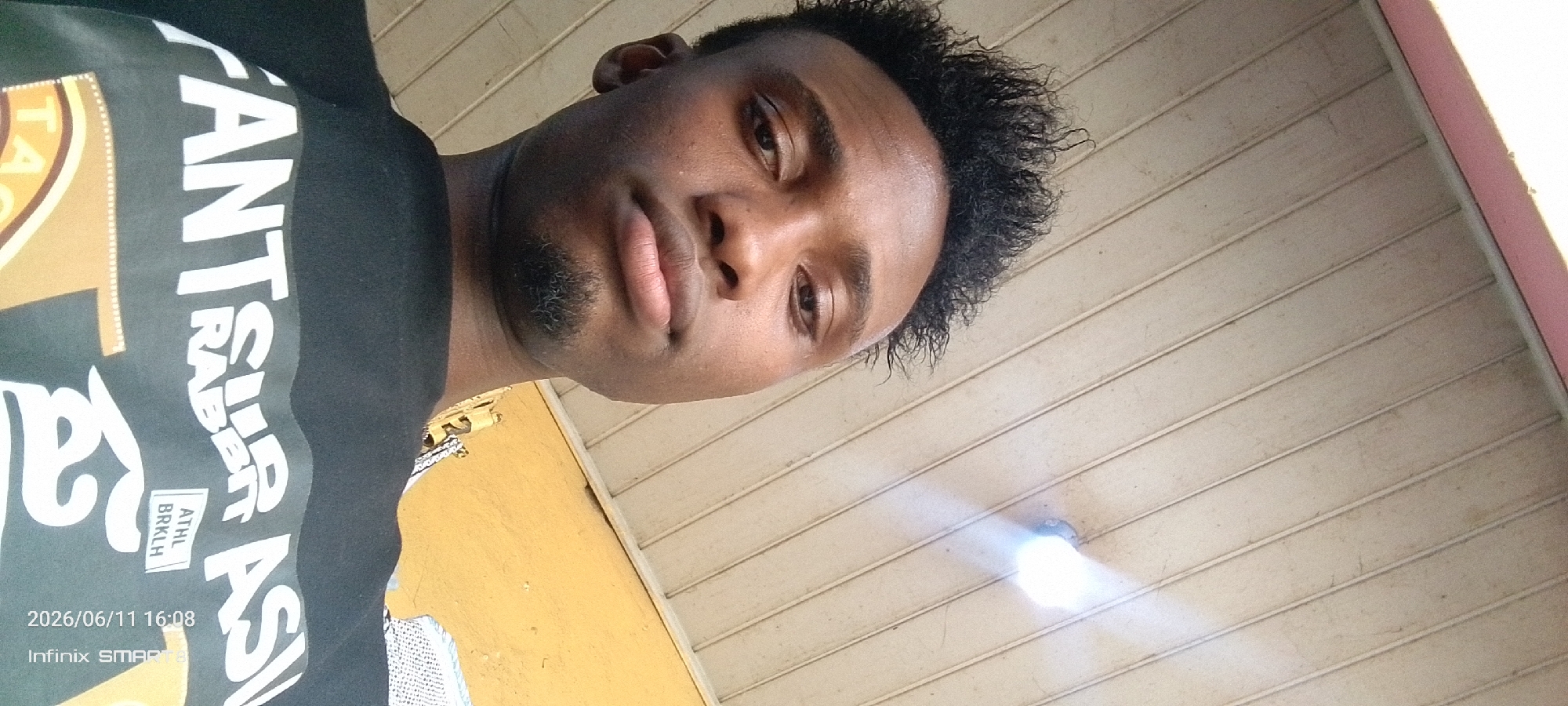 A close-up, tilted, outdoor shot shows a young Black male from the chest up, wearing a black t-shirt with white text. The t-shirt reads "YANTSURA RABBI" and "ATHL BRK LH." The male has dark skin, short, curly black hair, and a light beard. His face is turned towards the camera, with his eyes looking slightly to the left. The background consists of a yellow wall on the left and a beige-colored corrugated ceiling on the right. A bright light fixture is visible on the ceiling. The bottom left corner of the image displays a timestamp "2026/06/11 16:08" and "Infinix SMART 8."