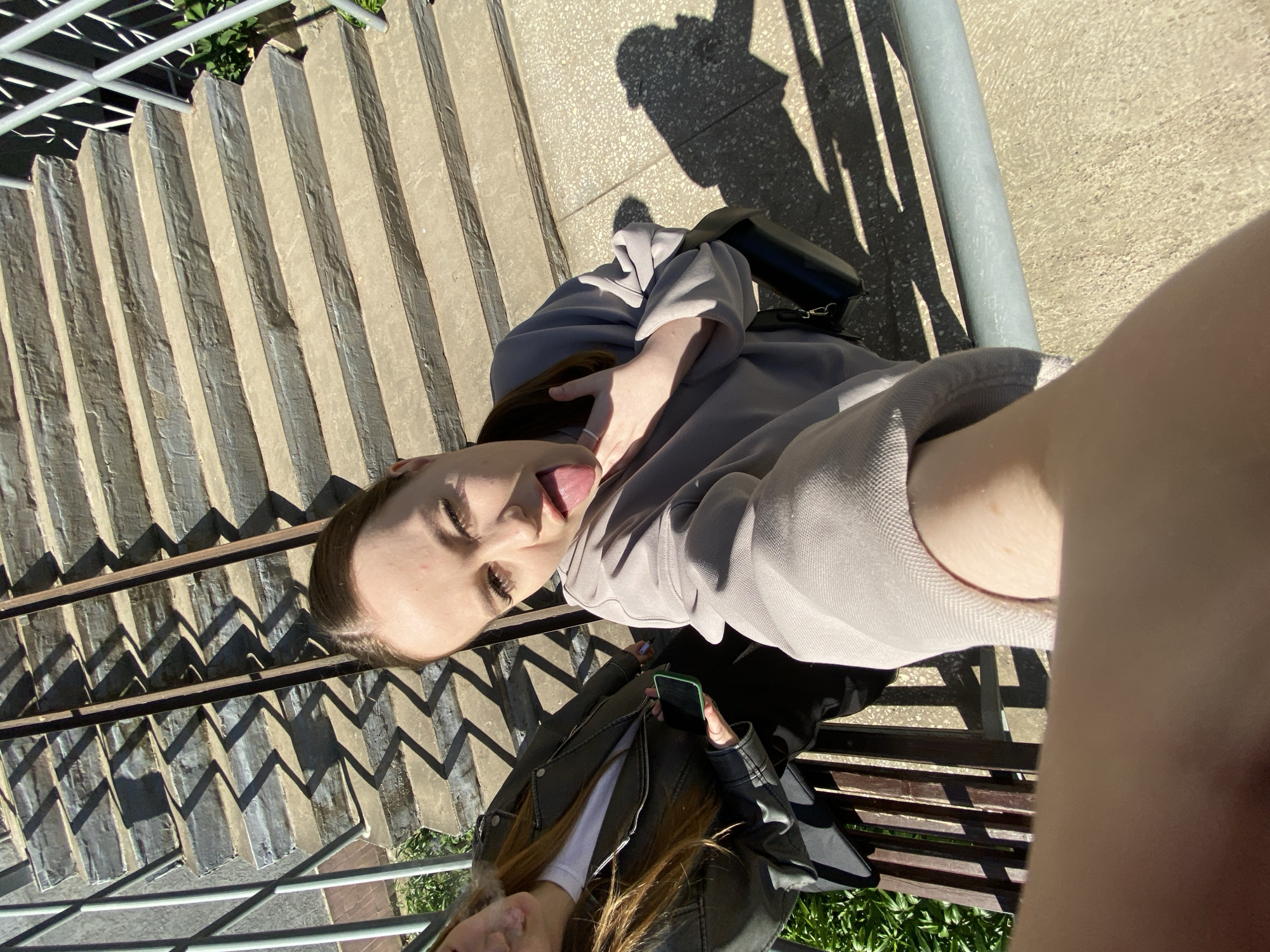 In Chaikovsky, Russia, two young women are captured in a selfie on a sunny day. They are seated on a wooden bench near a set of concrete stairs. The woman in the foreground is leaning forward, sticking her tongue out at the camera, with her right hand touching her face. She is wearing a light gray sweatshirt. Next to her, the second woman, on the left side of the frame, is also looking towards the camera. She has long, reddish-brown hair and is wearing a dark leather jacket over a white t-shirt. She is holding a black smartphone in her left hand. The background shows a shadowed pattern cast by a metal railing onto the stairs, and lush green foliage can be seen to the left and right of the stairs. The overall atmosphere is casual and relaxed.