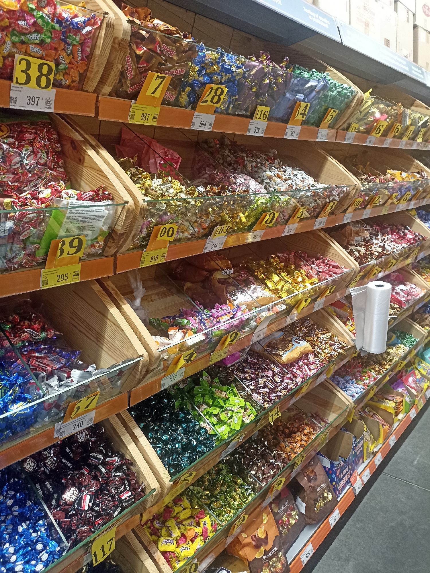 The image shows a retail display of bulk candies in a supermarket. The candies are arranged in wooden bins on shelves. Many of the bins have price tags indicating a number and a price in what appears to be Kyrgyzstani Som. The candies are individually wrapped in colorful foil and plastic packaging. Some of the visible numbers on the price tags are 38, 397, 7, 28, 116, 14, 15, 6, 100, 915, 39, 295, 57, 740, 117, 4, 64, 372, 18, 2, 6, 103, 344, 307, 539, 563, 113, 657, 1649, 356. The background includes other retail displays and what appears to be a ceiling with lighting. The image is well-lit and the focus is on the candy display. The scene is set in the city of Osh City, Kyrgyzstan.