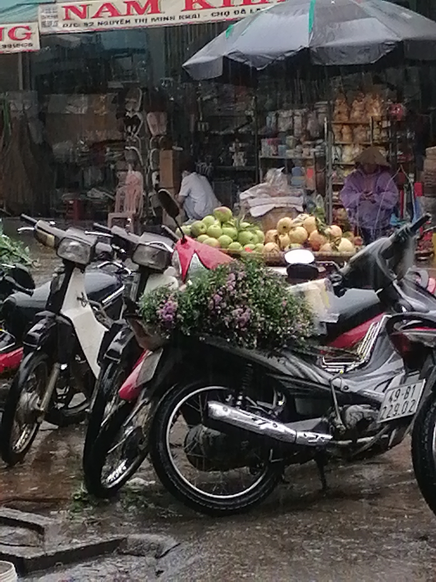 The image depicts a rainy day scene at a market stall in Da Lat, Vietnam. In the foreground, two parked motorcycles, one white and black, the other black with red accents, are visible. The white and black motorcycle has its headlight illuminated. A cluster of purple flowers is placed in a basket on the handlebars of the black motorcycle. The ground is wet and reflects the surroundings, indicating recent rainfall.
Behind the motorcycles, the market stall is set up with shelves displaying various goods. A person wearing a conical hat and a purple jacket stands near the stall, holding a mobile phone. To the left, a pile of fruits, possibly apples and pears, is displayed. In the background, a person in a white shirt is partially visible. A large black umbrella covers part of the stall. A sign with Vietnamese text, partially obscured by rain, is mounted above the stall. The overall atmosphere is that of a busy, wet marketplace.