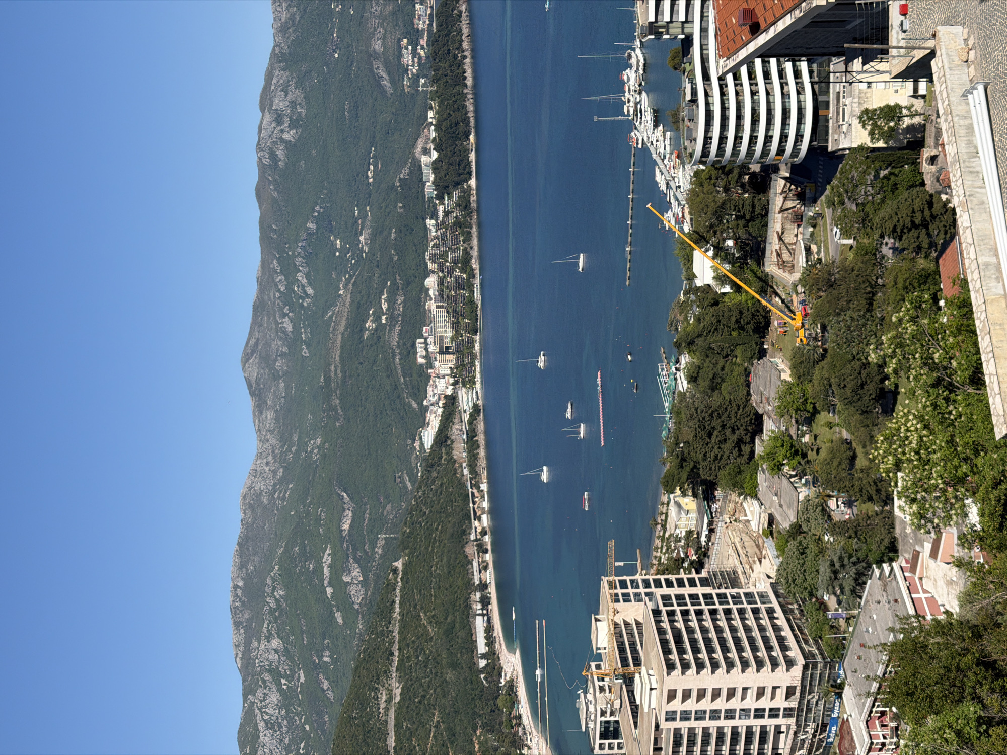 This is an aerial view of Budva, Montenegro, featuring a narrow body of water with several boats and a coastline with buildings nestled between the sea and a mountain range. Construction cranes are visible among the structures, and a modern white building with a curved facade is located near the center. Trees, buildings, and boats are scattered across the landscape.