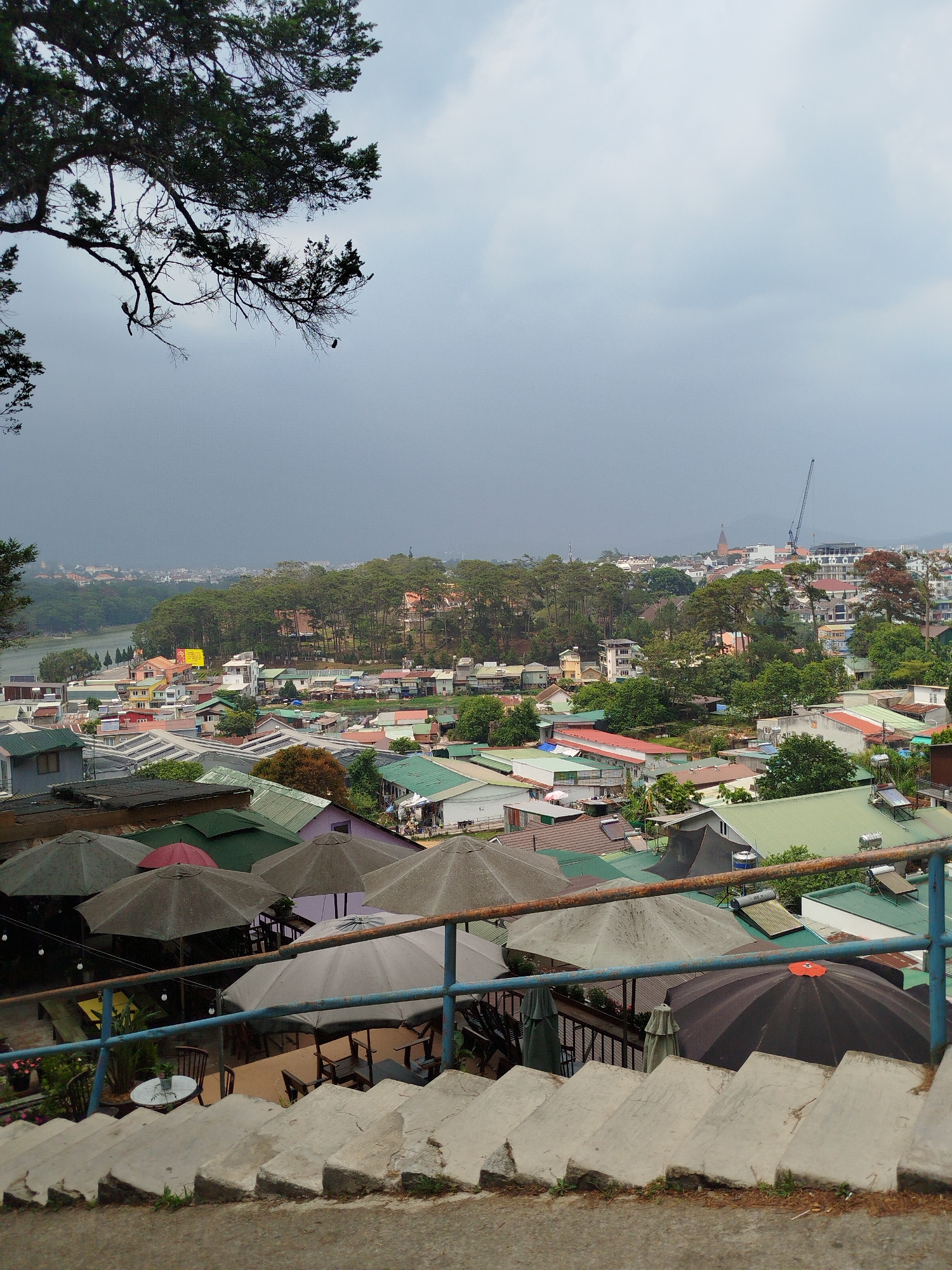 An elevated view of Da Lat, Vietnam, from a set of concrete stairs with a blue metal railing. Several gray and black umbrellas cover outdoor seating areas in the foreground. Buildings with multi-colored roofs, predominantly green and red, fill the middle ground. A dense line of trees is visible in the distance, with a river or lake situated on the left. In the background, buildings are visible in the cityscape, one with a spire and a construction crane. The sky is overcast with dark clouds suggesting possible rain. Part of a tree occupies the upper left corner.
