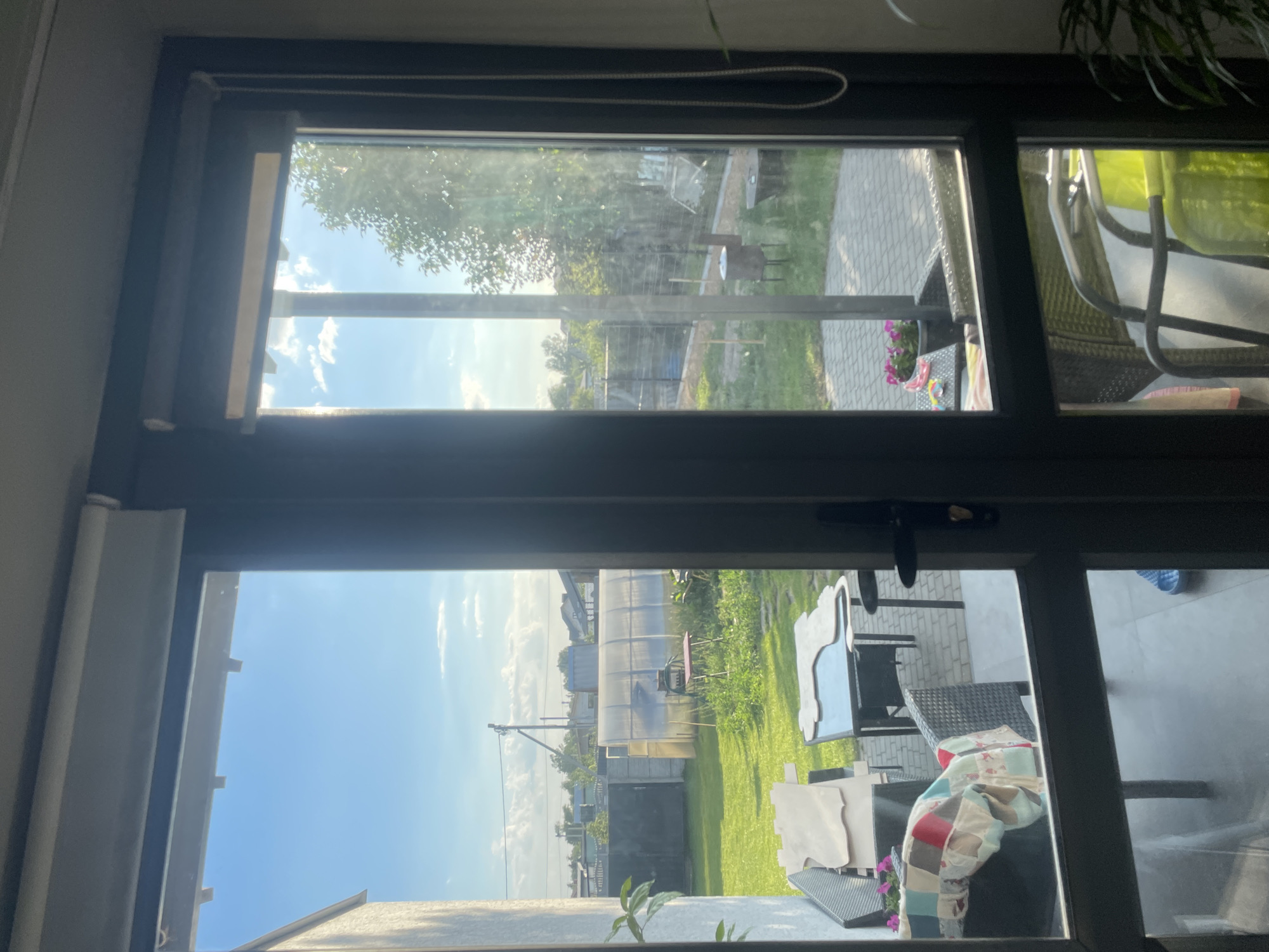 This media file is a photograph taken from inside a room, looking out through a large window. The window appears to be divided into multiple panes.
The view outside is a sunny, outdoor scene. In the upper pane, sunlight filters through green trees, casting dappled shadows on a paved patio area. A glimpse of outdoor furniture, including a chair with a bright green cushion, is visible. A small pot of pink flowers sits on a table. The sky above is blue with scattered white clouds.
The lower pane of the window shows a backyard with a neatly mown green lawn. A large, translucent greenhouse or polytunnel dominates the background, with smaller outbuildings and houses visible beyond it. A picnic table with chairs is situated near the greenhouse. There are also stacks of what appear to be building materials or large decorative items on the right side of the frame. The sky in this section is a bright, clear blue, suggesting a pleasant day.
The overall impression is one of a bright, sunny day in a residential or garden setting. The time of day appears to be late morning or early afternoon, based on the angle of the sunlight. There are no people visible in the scene. The setting seems to be rural or suburban, possibly in a region with a temperate climate. The photograph has a slightly hazy quality, which could be due to the window glass or atmospheric conditions. There is no visible text in the image.