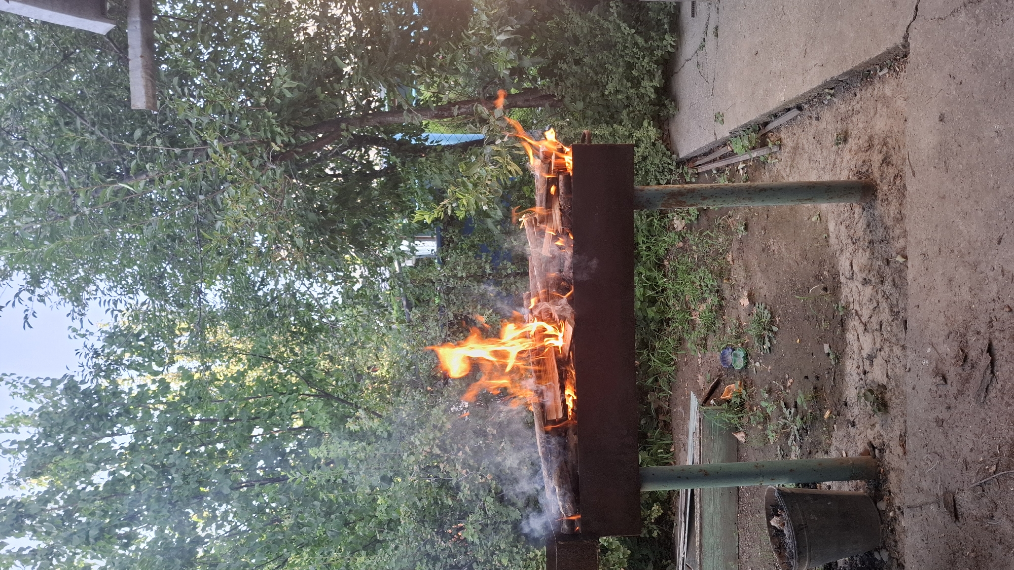 In the city of Shubarssu, Kazakhstan, a metal barbecue grill is set ablaze with a fire. The grill, supported by two teal-colored metal legs, is filled with burning logs that produce bright orange flames and wisps of smoke. The surrounding environment is a courtyard with a dirt floor and scattered grass. To the left of the grill, a dark metal bucket is visible. In the background, lush green trees and foliage create a natural backdrop, suggesting an outdoor setting. A concrete pathway is visible on the right side of the image, with visible cracks. The sky above is a clear, light blue. There are no people present in the image.