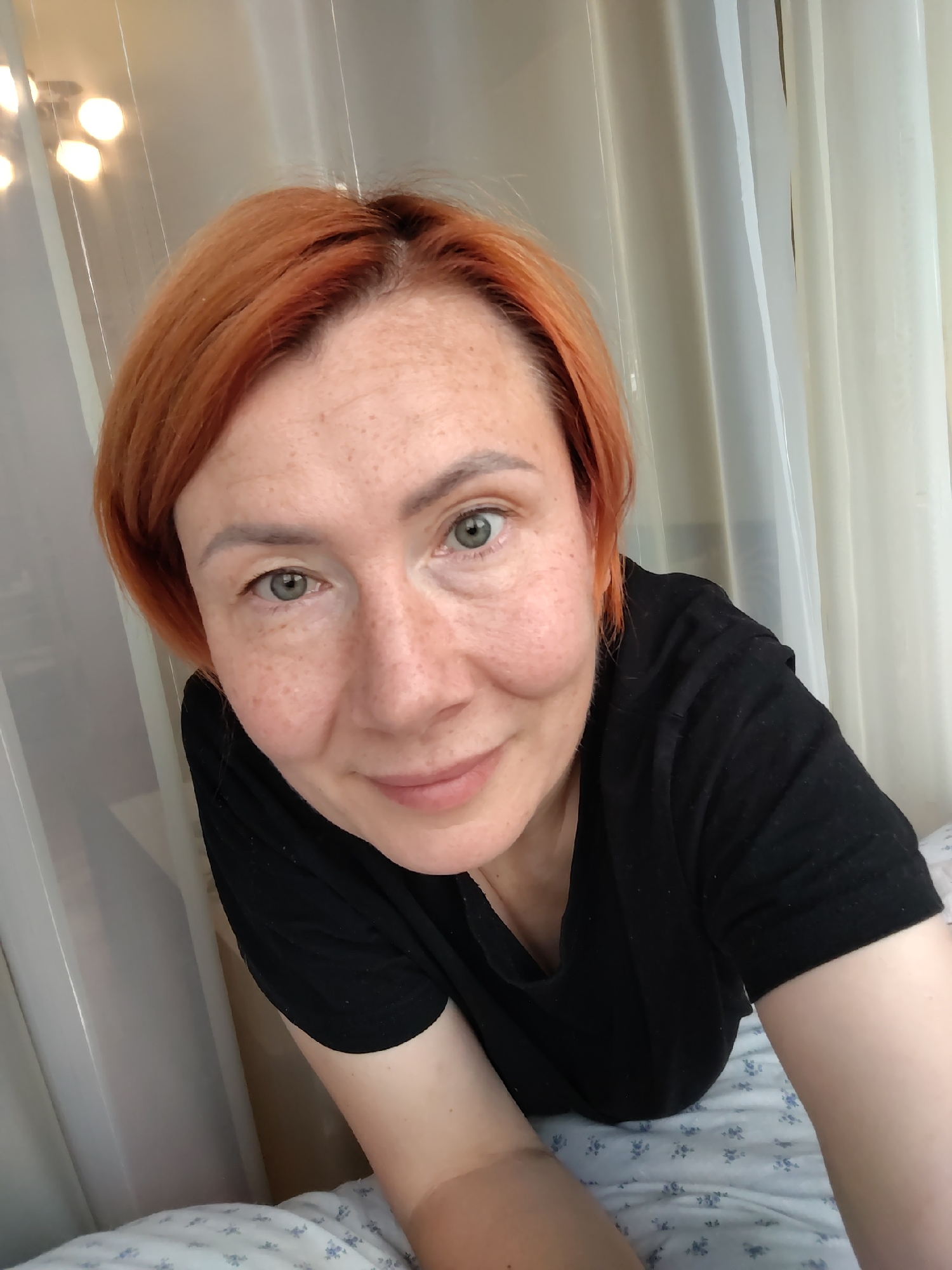 A close-up, eye-level shot features a woman with reddish-orange hair and fair skin with freckles. She is wearing a black t-shirt and lying on a bed with a floral patterned duvet. The background consists of white curtains and some out-of-focus lights. The image does not contain any identifiable landmarks or contextual information that would allow for its location to be specified within the city of Konakovo, Russia.