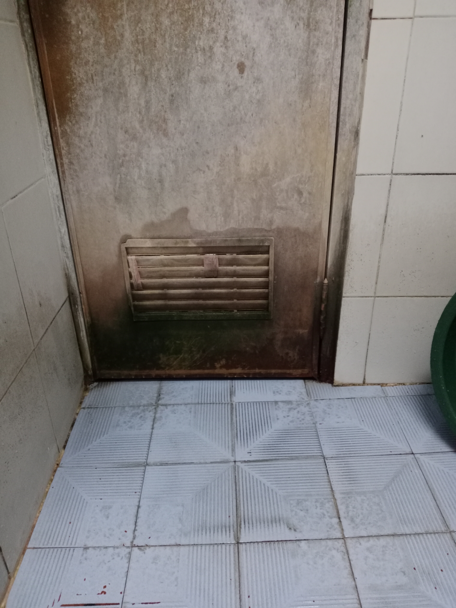 The image depicts a corner of a room with tiled walls and floor, featuring a metal door with a vent. The environment appears somewhat neglected, with visible signs of dirt and discoloration on the door and wall tiles. The floor is tiled with a pattern of raised lines. The lighting suggests an indoor setting, possibly a utility area or bathroom. There are no people or active events depicted in the image. The overall impression is one of a mundane, possibly damp, interior space.