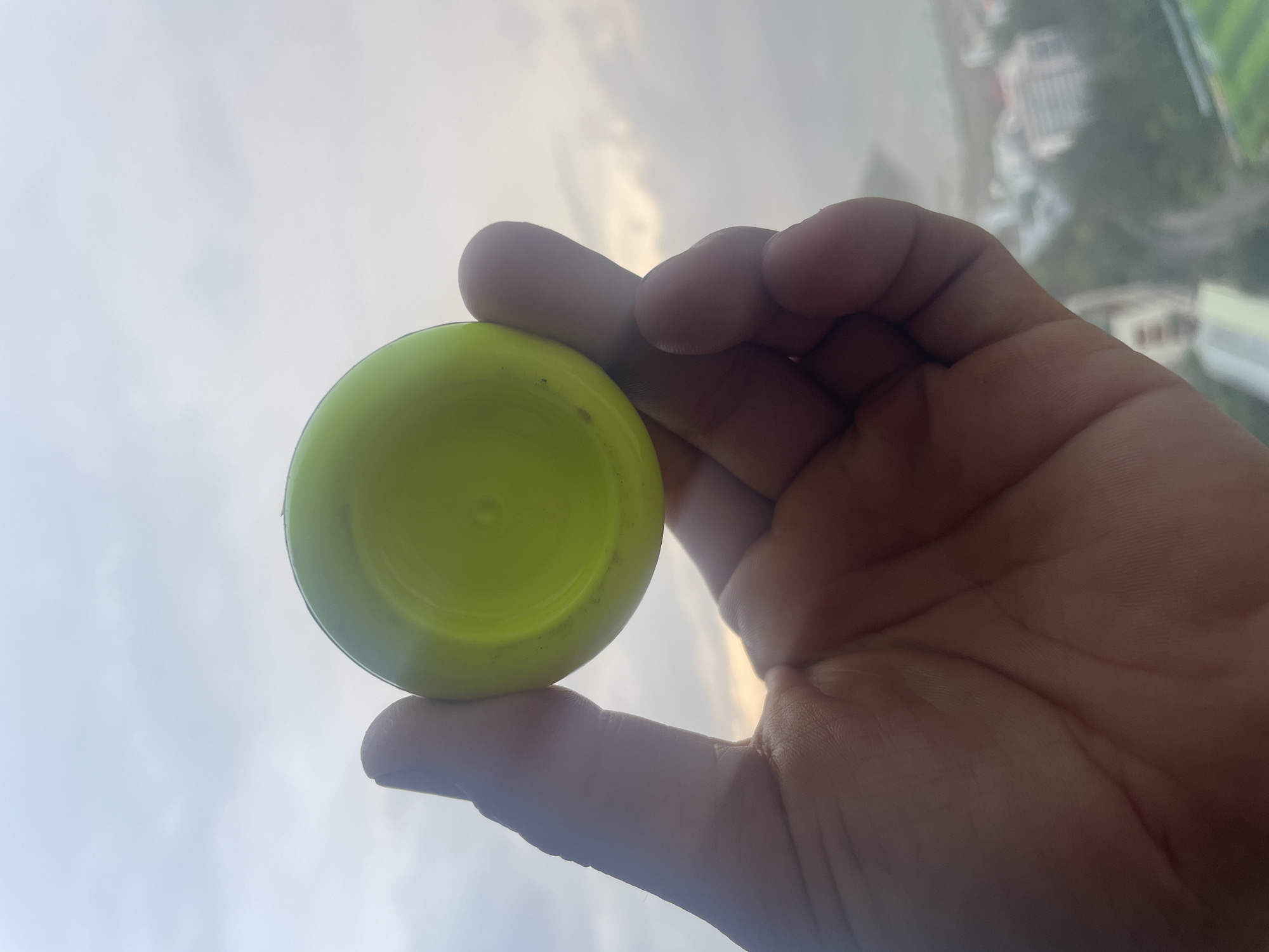 A hand holds a yellow plastic lid against a background of cloudy sky and buildings. The location appears to be in or near Bac Nha Trang Ward, Vietnam. The buildings are blurred due to distance and focus. The hand is fair-skinned and appears to be holding the lid between the thumb and index finger.