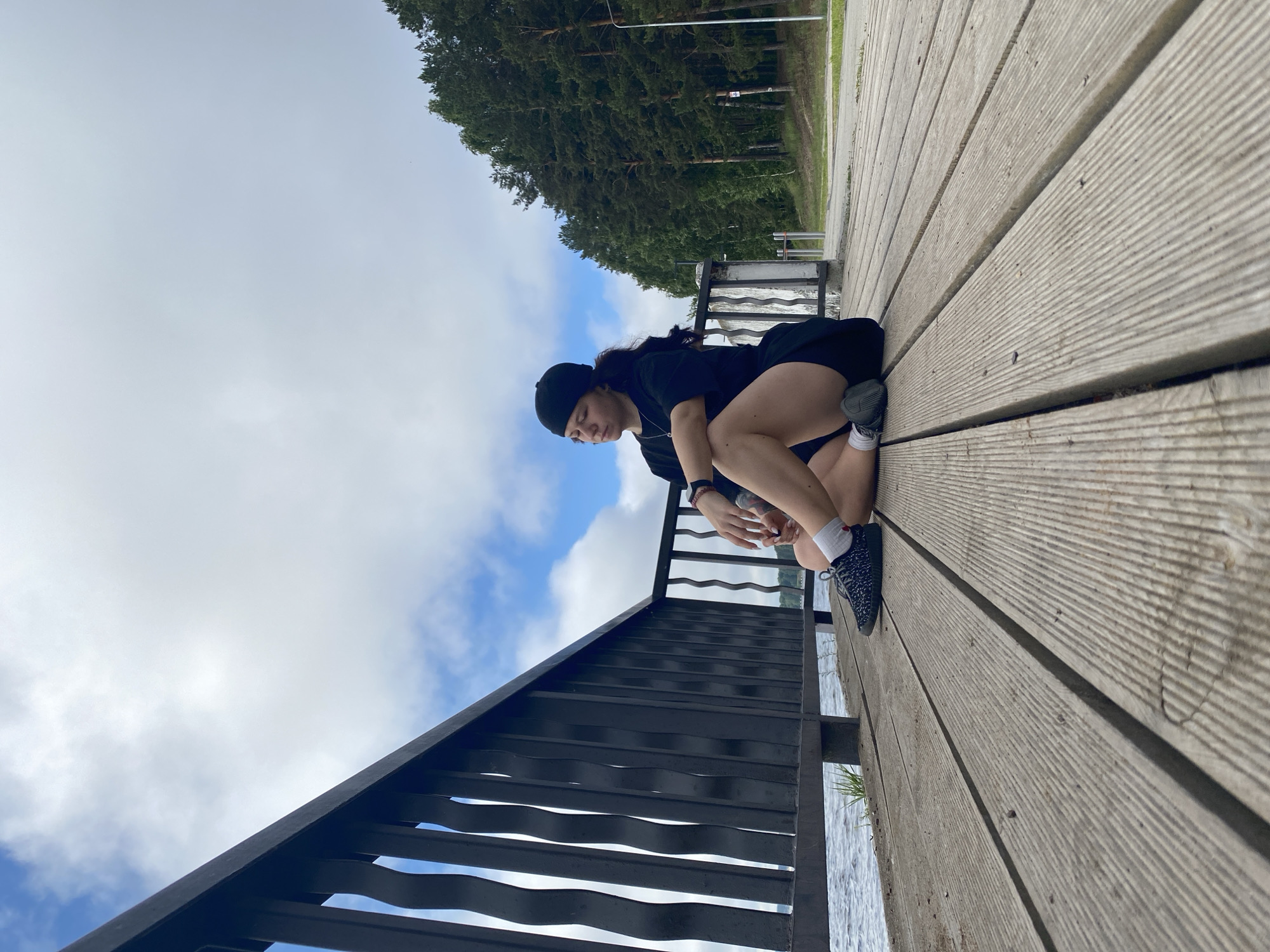 A young woman with dark hair pulled back in a ponytail, wearing a black t-shirt, black shorts, a black beanie, and black and white sneakers, sits cross-legged on a wooden pier. She is positioned to the right of the frame, facing left, with her eyes closed and her head tilted back slightly. A black metal railing runs along the left side of the pier, separating it from the choppy water. Beyond the railing, the water stretches out towards a distant tree line. The pier is constructed of weathered wooden planks. The sky above is a mix of white clouds and patches of blue. The background includes a dense forest of green pine trees and a paved path. The location is the embankment of the Kama River in Chaikovsky, Russia.