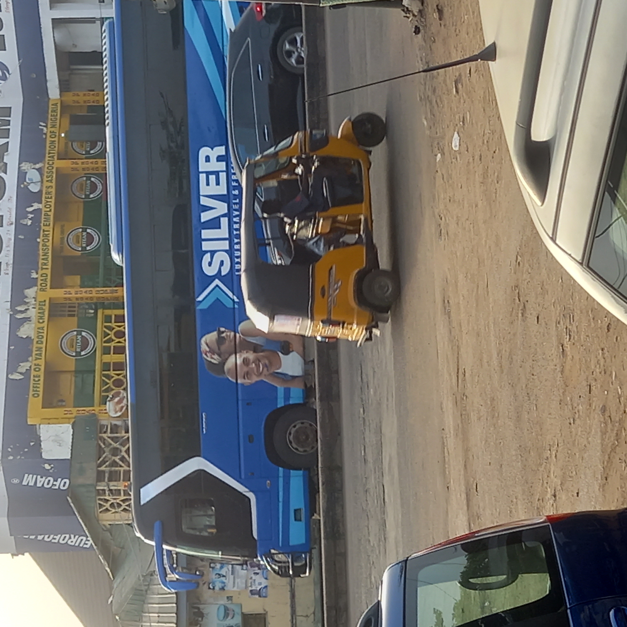 The image captures a bustling street scene during daylight, likely in Dadi Riba, Nigeria, given the visible text and vehicle types. The main subjects are a large blue bus, a yellow motorized tricycle (often called a 'keke' in Nigeria), and several cars, along with two men smiling from inside the bus.
The blue bus, branded "SILVER LUXURY TRAVEL & FR" (likely 'Freight' or 'Facilities'), dominates the left side of the frame. Two men are prominently visible through a window of the bus, one bald and the other wearing a patterned headscarf or bandana, both smiling. To the right of the bus, a yellow tricycle with a black canopy is present, seemingly on the road or roadside, with a driver visible inside. Further right, parts of other cars, including a dark sedan and a white car, are visible, suggesting traffic or parked vehicles. The road surface is unpaved or dirt-covered in places.
In the background, behind the blue bus, stands a building with yellow and green sections, displaying several signs. Visible text on this building includes "OFFICE OF YAN DOYA CHAPEL ROAD TRANSPORT EMPLOYER'S ASSOCIATION OF NIGERIA", and repeated instances of "RTEAN" and "UP ROAD". Additionally, "EUROFOAM" is visible on a sign attached to an unseen structure to the far left. The overall scene conveys a lively, everyday moment in an urban or semi-urban setting.