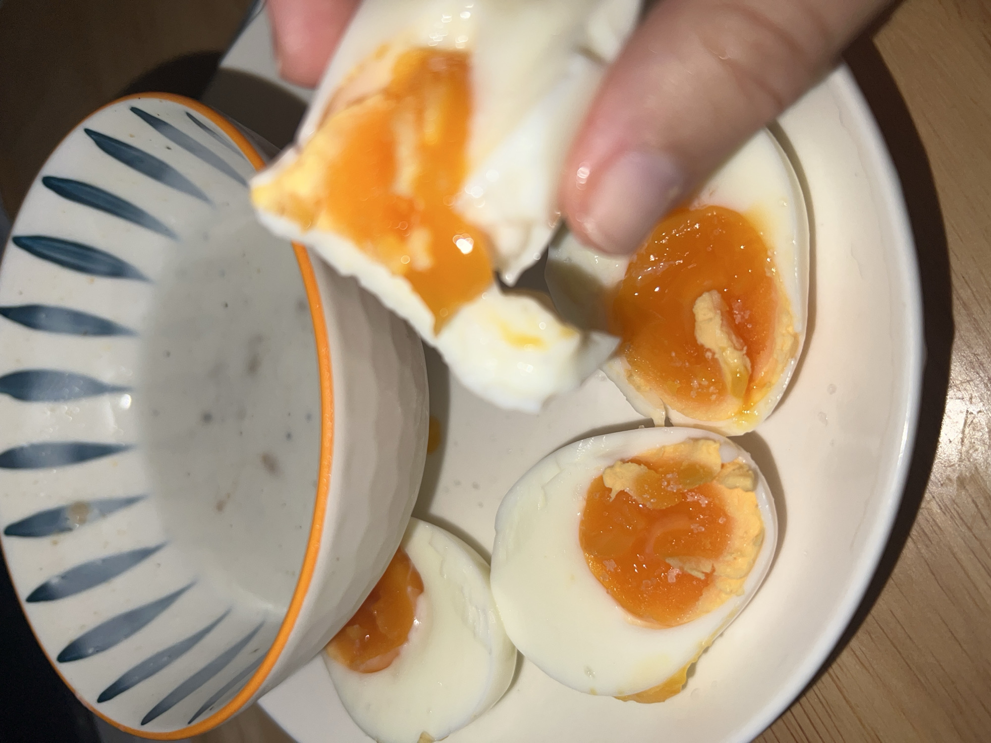 A close-up shot captures a hand holding a piece of a soft-boiled egg, revealing a bright orange, runny yolk. On a white ceramic plate, several other soft-boiled egg halves are visible, each showcasing a vibrant, semi-liquid yolk contrasting with the firm white albumen. Small grains, likely salt, are sprinkled on the surface of some yolks. To the left, a ceramic bowl with a distinctive blue-grey striped pattern and an orange rim is partially visible, containing a light greyish liquid, possibly a sauce or a thin soup. The meal is presented on a wooden surface, suggesting an indoor dining setting with artificial lighting. This meal featuring soft-boiled eggs is being consumed in the Nam Natural Heritage Area Trang, Vietnam. No text is visible in the image.