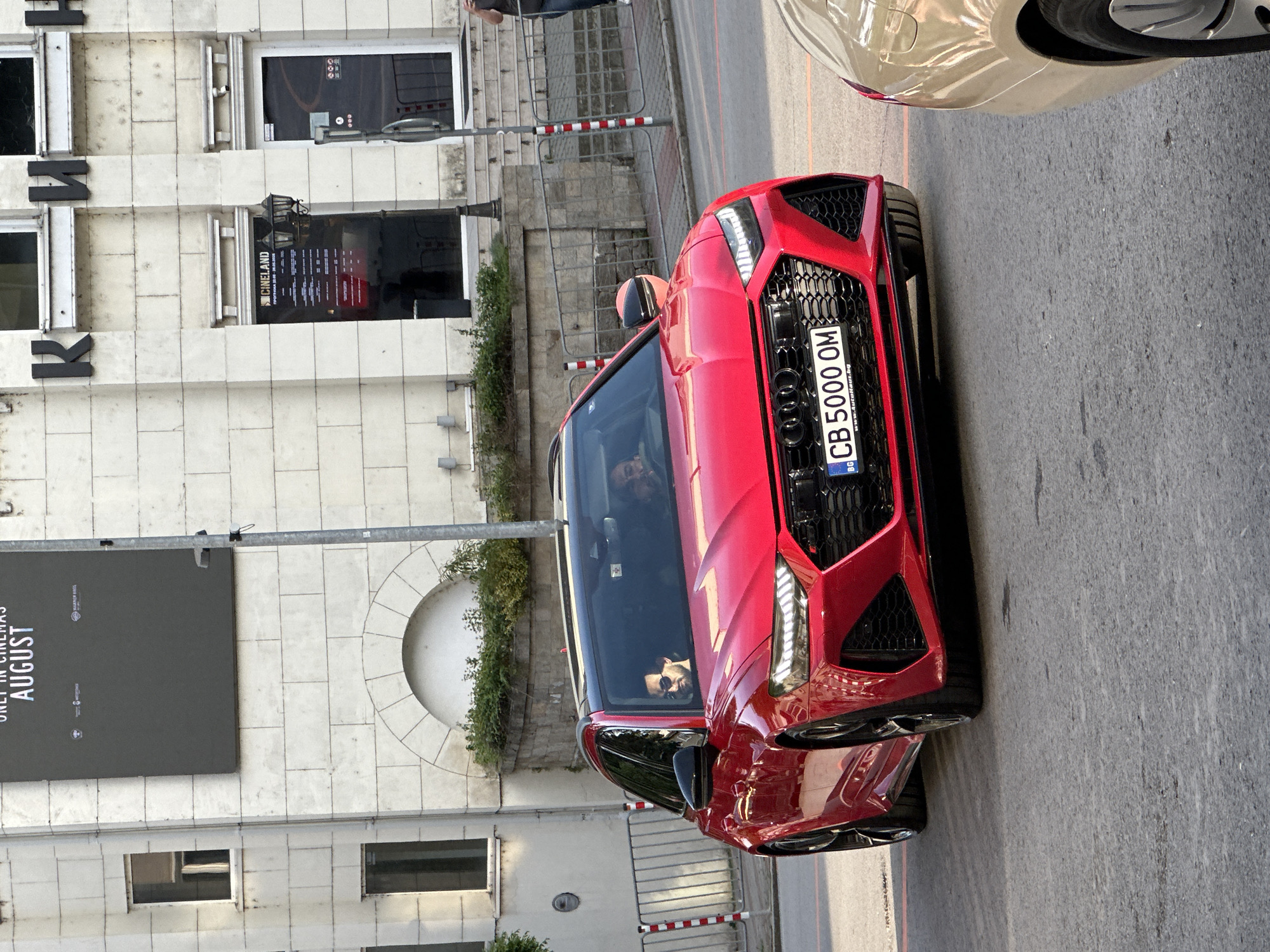 A red Audi RS6 Avant, with Bulgarian license plate CB 5000 OM, is parked on a street in Veliko Tarnovo, Bulgaria. A driver is visible inside the vehicle. The car is in front of a building with a sign that reads "KINOLAND", adjacent to a red-and-white striped pedestrian barrier. A black billboard for a movie with "AUGUST" printed on it is also visible.