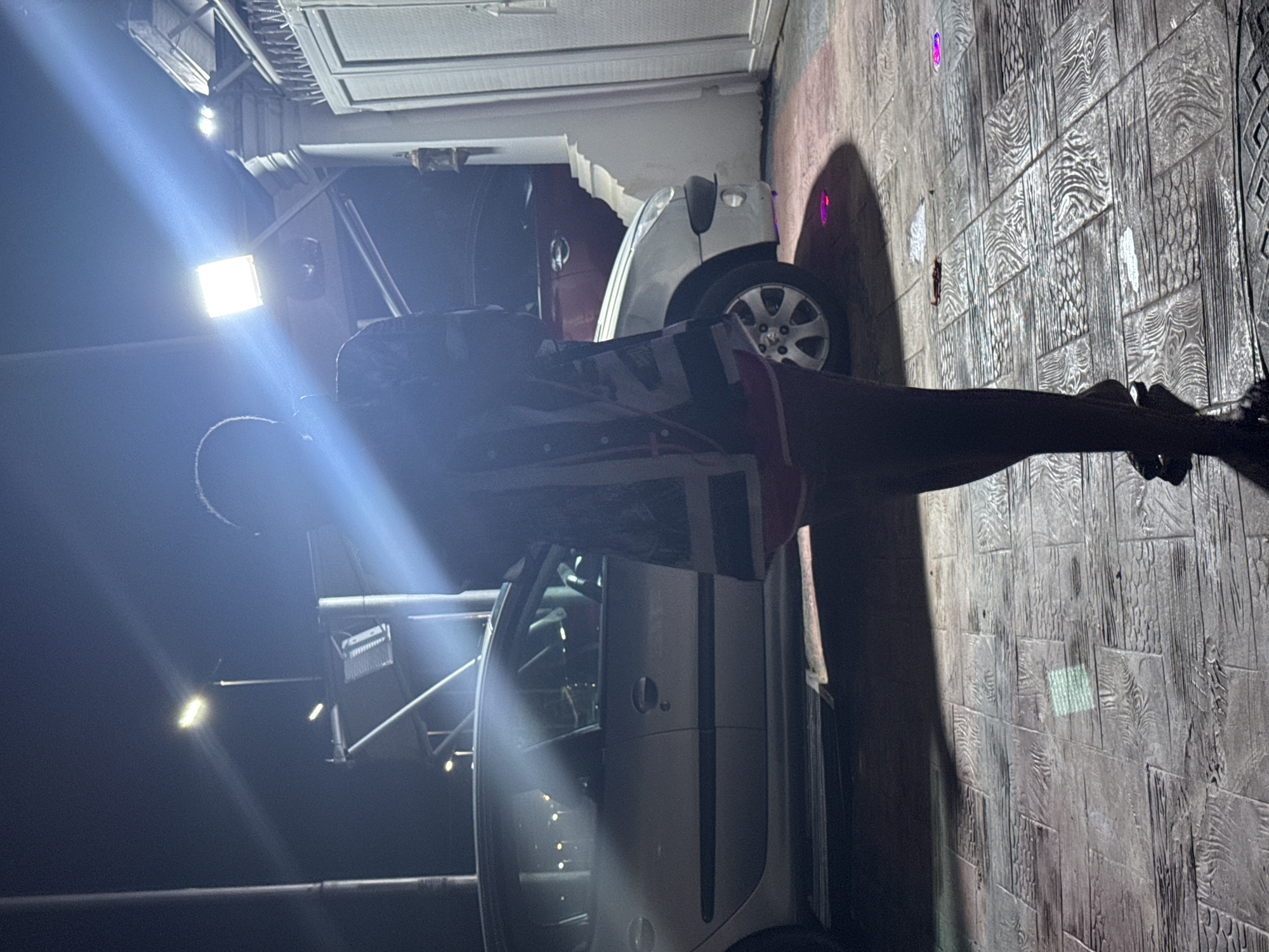 The image depicts a nighttime scene in Abuja, Nigeria, where a person stands silhouetted against a bright light source. The person, facing away from the camera, is wearing a patterned shirt and shorts, and appears to be standing on a patterned tiled ground. Behind the person, a silver car is parked. In the background, there is a gate and a white structure, possibly part of a building. The lighting suggests an outdoor setting, potentially a driveway or a courtyard, with artificial lights illuminating the area. The presence of cars and the architectural elements hint at an urban or suburban environment.