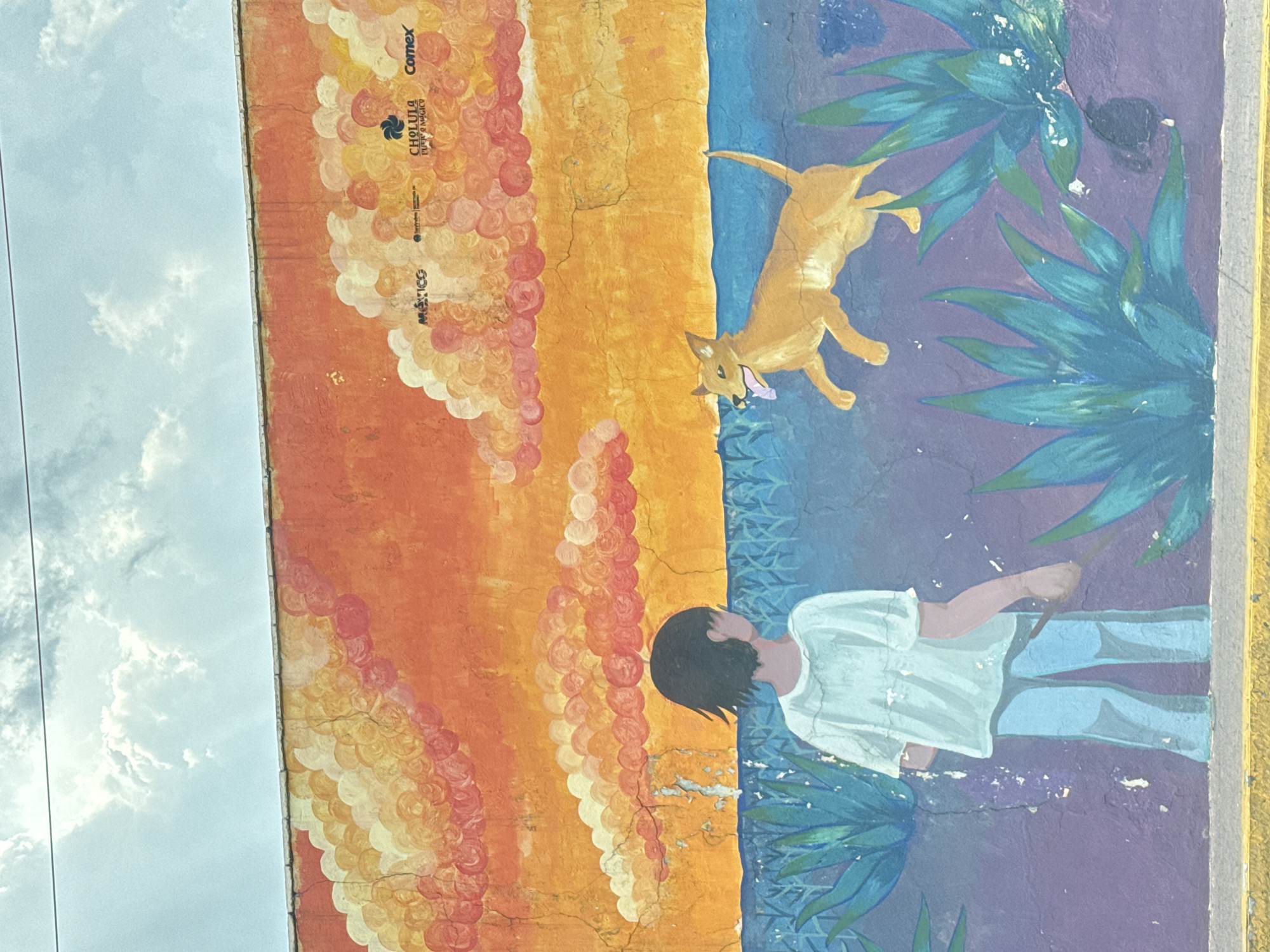 The image is a mural on a building located in San Andrés Cholula, Mexico. The mural is divided into two distinct color palettes. The left side features an orange and yellow gradient backdrop with numerous round, cloud-like shapes. The words "México," "Cholula Pueblo Mágico," and "Comex" are printed on the upper left side of the mural. The right side features a blue and purple backdrop. A tan dog is depicted jumping in the upper right area, while a person is depicted below in a white shirt and light blue pants. Both are surrounded by green plants. The mural appears to be painted on a rough, slightly cracked surface, and the photograph of the mural also captures a portion of the sky and telephone wires in the background.