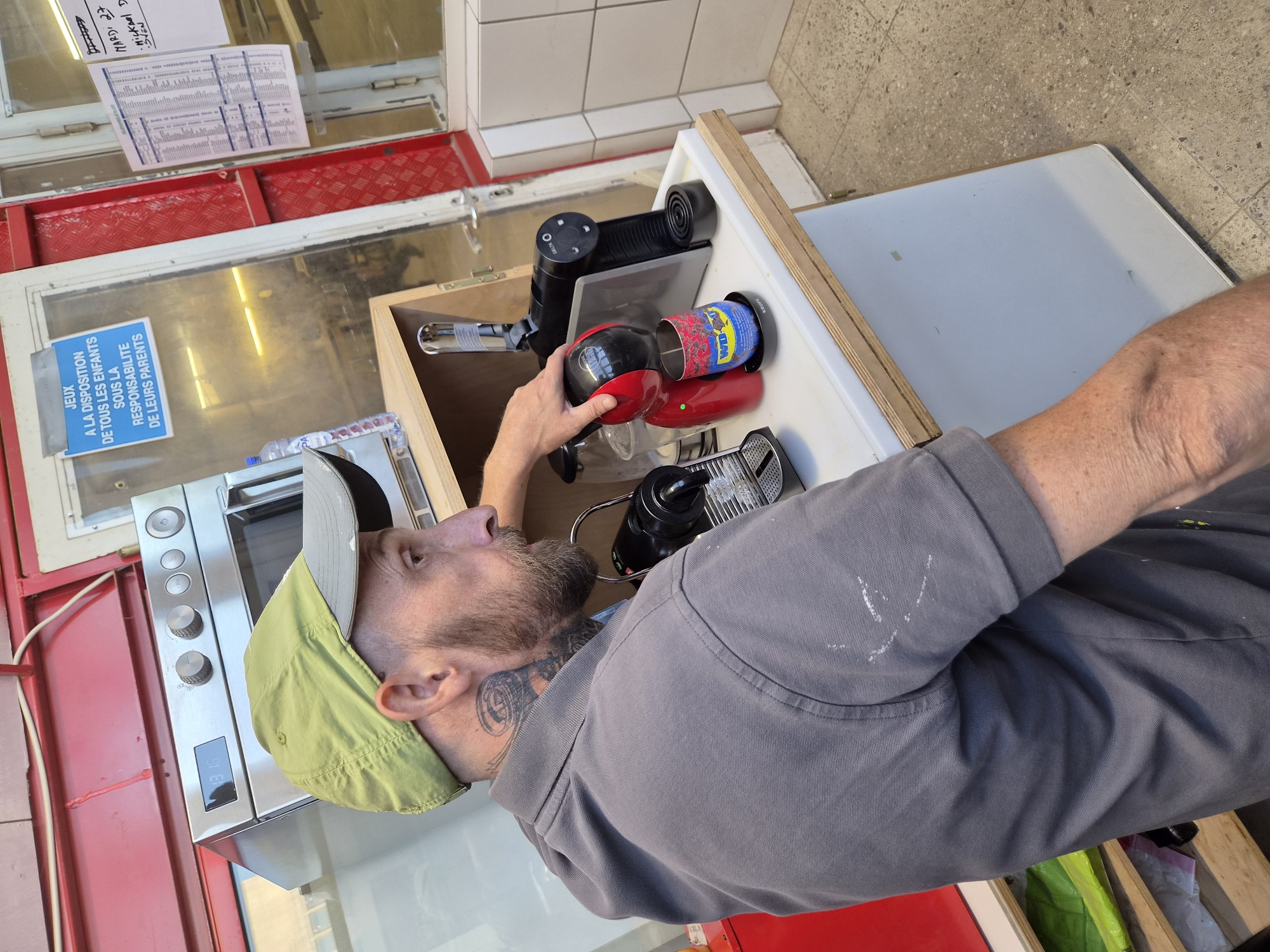 A man with a beard and neck tattoos wearing a grey polo shirt and a green cap is standing in what appears to be a breakroom or utility area, possibly in Leudelange, Luxembourg. He is reaching for a red and black coffee machine, possibly a Dolce Gusto. To his right is a white counter with a white tiled wall behind it, and to his left is a stainless steel oven. A blue sign with white lettering is visible on a glass door in the background, reading "JEUX A LA DISPOSITION DE TOUS LES ENFANTS SOUS LA RESPONSABILITE DE LEURS PARENTS" (Toys available to all children under the responsibility of their parents). A white sheet of paper with handwritten text is attached to a red metal support beam to the right of the glass door. The floor is tiled with a grey speckled pattern.