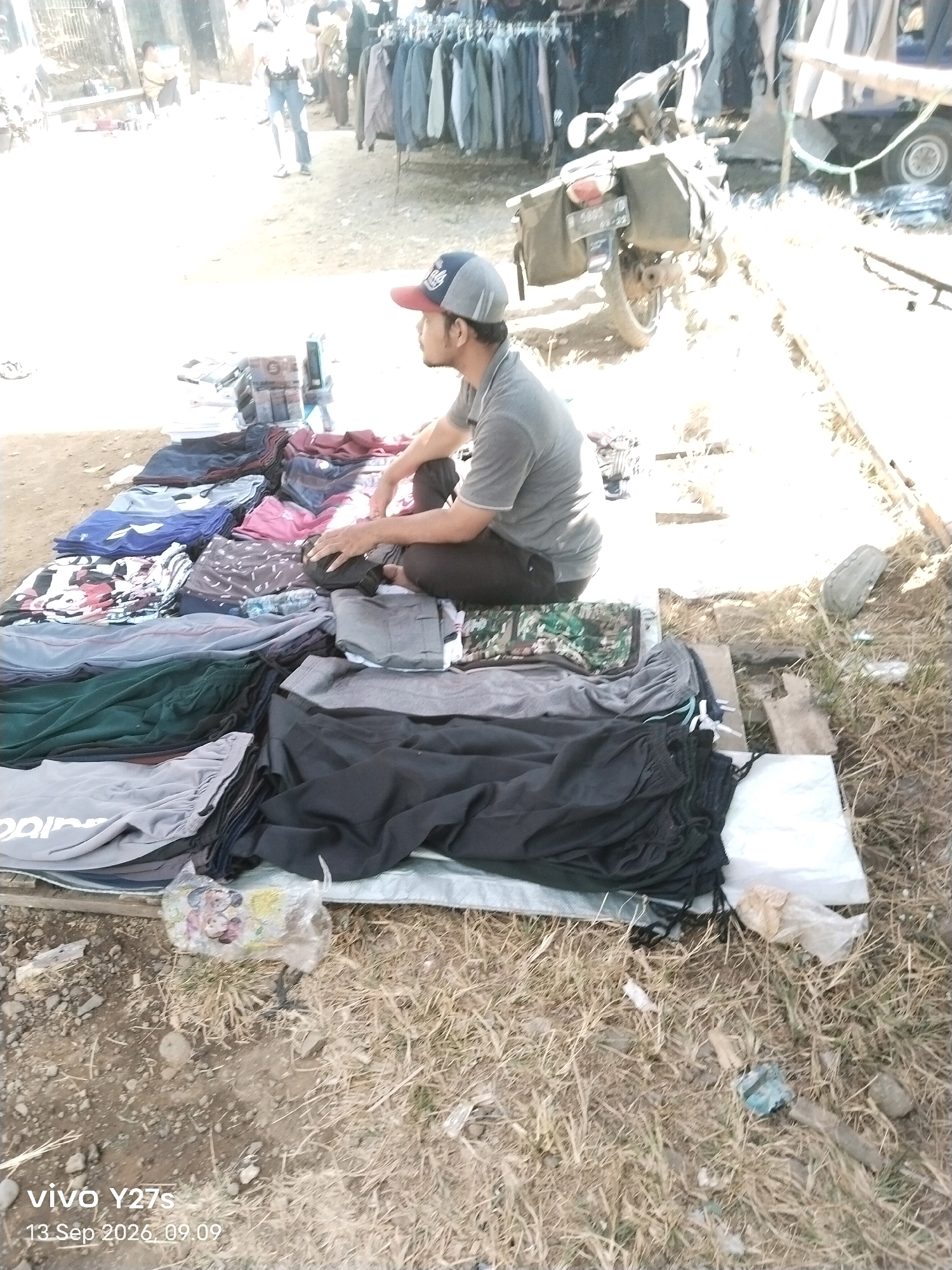 The image depicts an outdoor market scene in Trimulyo, Indonesia, likely a roadside stall. A man wearing a gray t-shirt and a red and gray baseball cap is seated on the ground amongst a collection of folded clothing, primarily pants and shorts. The clothing is neatly stacked on what appears to be a blue tarp. In the background, there are racks of hanging clothes, suggesting other vendors or a larger market area. A motorcycle with saddlebags is parked to the right of the central subject, and a glimpse of a cart or stall with wheels is visible. The ground is unpaved and appears to be a mix of dirt and dry grass. The overall lighting suggests it is daytime, with some harsh sunlight creating bright areas in the image. There are other indistinct figures and structures in the far background, hinting at a bustling market environment. The timestamp "vivo Y27s 13 Sep 2026 09.09" is superimposed on the bottom left corner of the image.