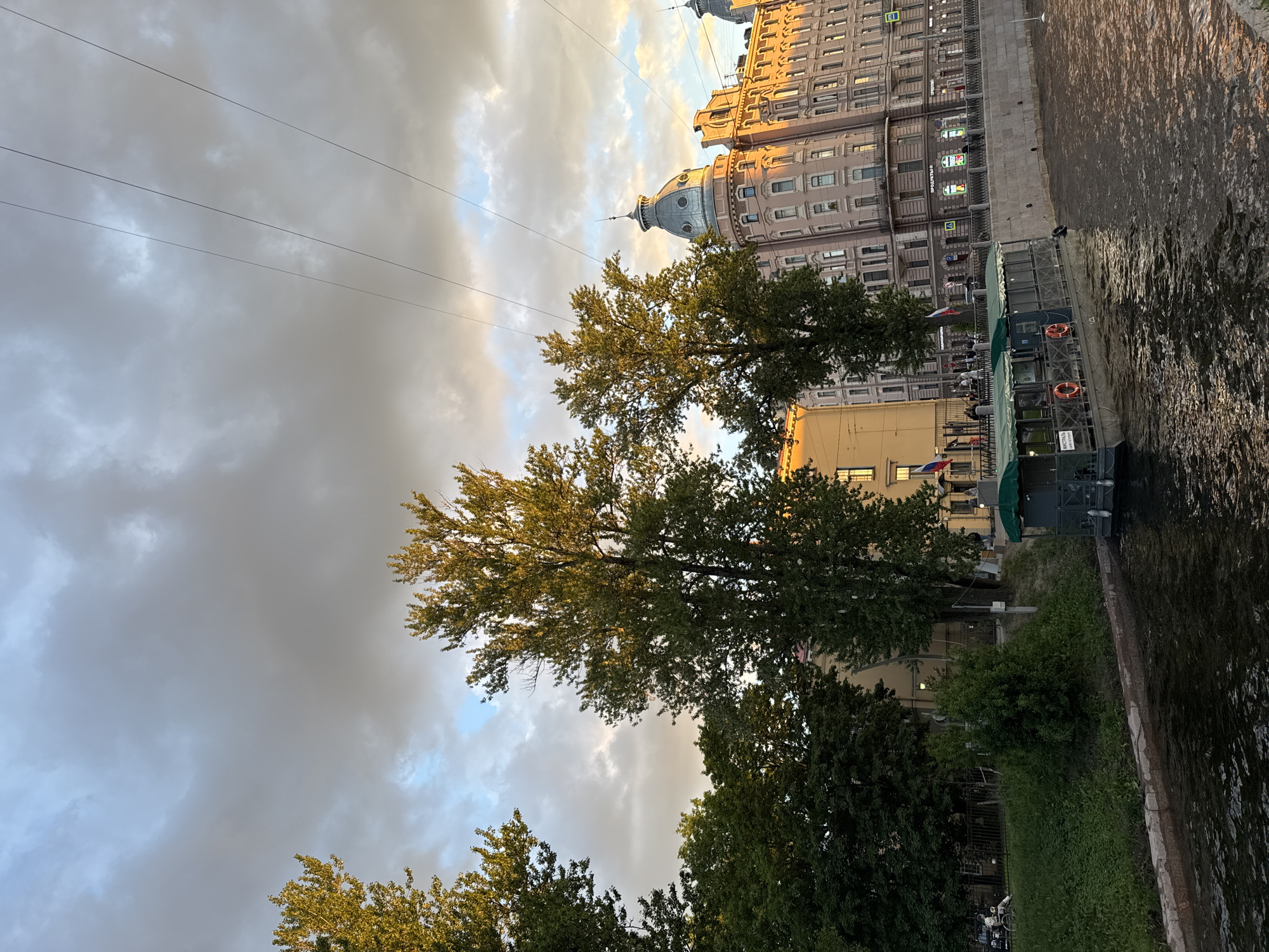 The image depicts a view of a canal in Saint Petersburg, Russia, likely during the late afternoon or early evening, given the warm, golden light illuminating the buildings. A tour boat with a green canopy is moored to the right side of the canal, its reflection shimmering on the dark water. Several people are visible on the boat, and a few are seated on the grassy bank to the left, where a person in a wheelchair is also present. The canal is lined by a stone embankment, behind which stands a row of multi-story buildings. The architecture suggests a historic district, with one prominent building featuring a rounded dome and ornate detailing. Tall trees with vibrant green foliage are scattered throughout the scene, with some branches reaching towards the sky. The sky above is a mix of dramatic, dark clouds and patches of lighter blue, with thin power lines crisscrossing the frame. Russian flags are visible on poles along the embankment.