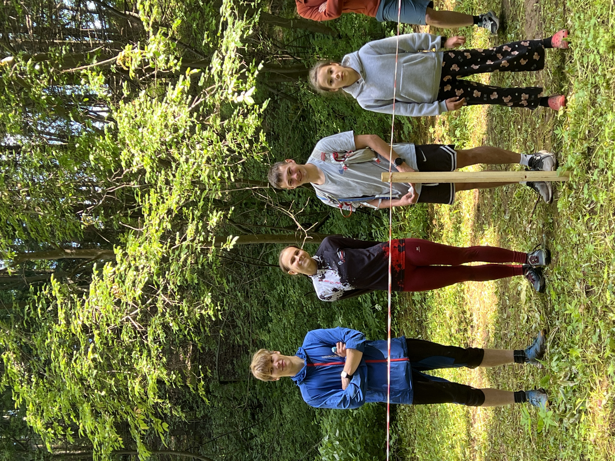 Four individuals, three young males and one young female, are standing in a wooded area. They are positioned behind a red and white striped tape that runs horizontally across the frame, acting as a boundary. The individuals are dressed in athletic attire. The person on the far left is wearing a blue hooded jacket, dark pants, and athletic shoes. The second person from the left is a female wearing a patterned long-sleeved shirt with black and white accents, dark red leggings, and athletic shoes. The third person from the left is a male wearing a grey t-shirt, black shorts, and athletic shoes; he is also wearing a hydration pack. The person on the far right is a young female wearing a grey hooded sweatshirt and patterned leggings with a pinkish hue, paired with pink footwear. The background consists of dense green foliage and trees, indicating a natural, possibly park or forest setting. Sunlight filters through the leaves, creating dappled light on the ground. The ground is covered with grass and fallen leaves. The overall scene suggests an outdoor event or activity taking place in a natural environment.