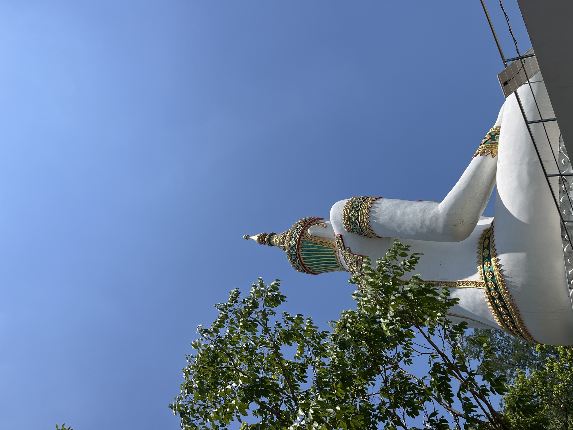The image shows a reclining white Buddha statue, adorned with gold and green ornamentation, viewed from a low angle. The statue is positioned against a clear, blue sky. In the foreground, branches and leaves of a tree partially obscure the view of the statue. To the right of the frame, portions of a building structure are visible. Based on the location metadata, the image likely depicts a religious site or temple in Nong Yaeng, Thailand.