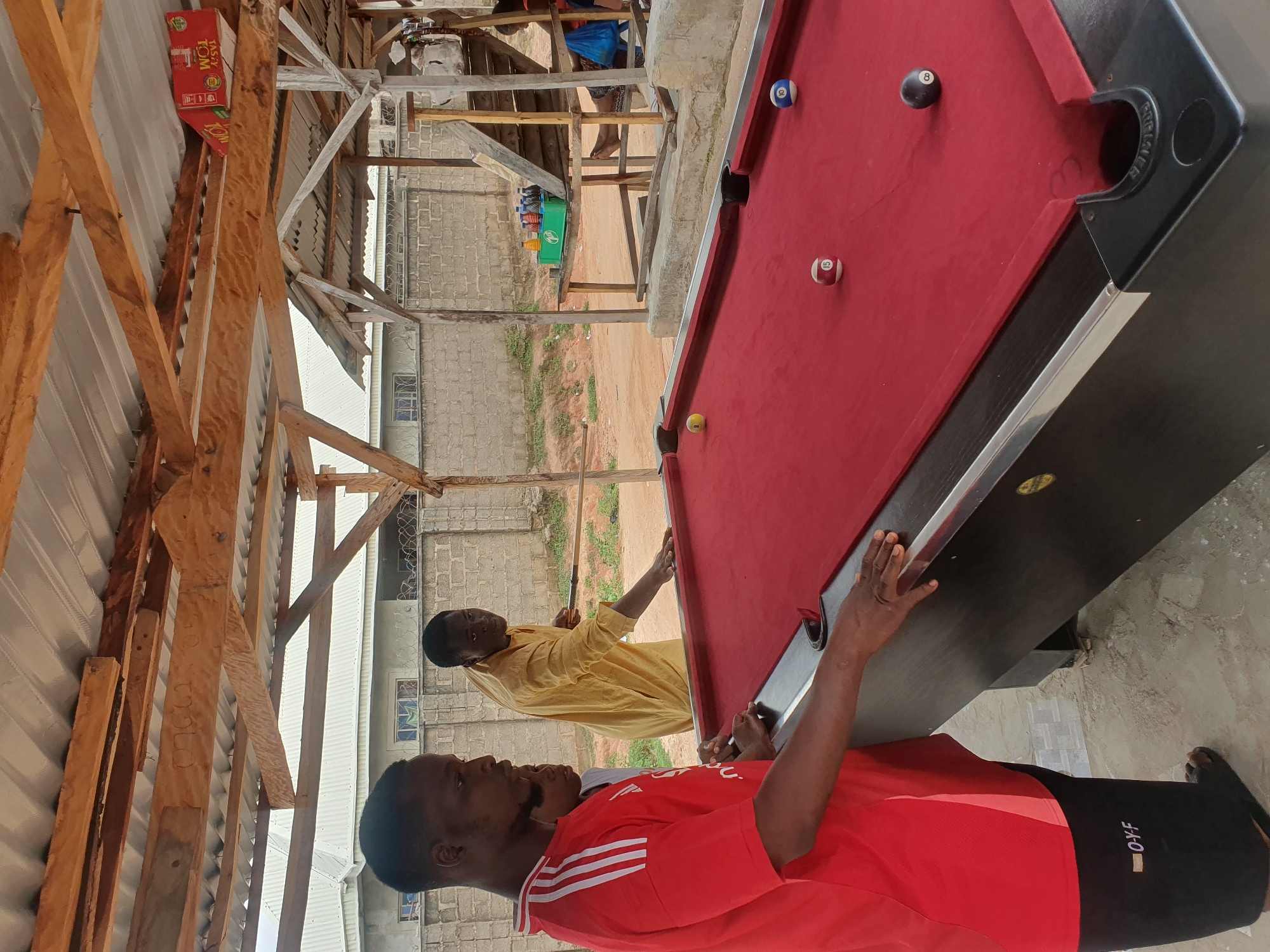 In Lokoja, Nigeria, three men are gathered around a red-felt billiard table under a corrugated metal roof supported by wooden beams. The man on the left, wearing a red shirt with white stripes and black shorts, is leaning on the table, his right hand resting on the edge. To his right, a second man, partially visible and wearing a grey shirt, stands by the table. The third man, in a yellow traditional garment, stands behind the table, holding a billiard cue and appearing to be playing. Several billiard balls are scattered on the table: a yellow ball on the left, a ball with the number 15 on the right, a ball with the number 8 in the center, and a ball with the number 10 on the far right. In the background, a brick wall with barbed wire runs horizontally, and beyond that, buildings with white roofs are visible. A wooden staircase leads up to an elevated platform on the right, where drinks in a green crate are placed. The ground is dirt and loose gravel.