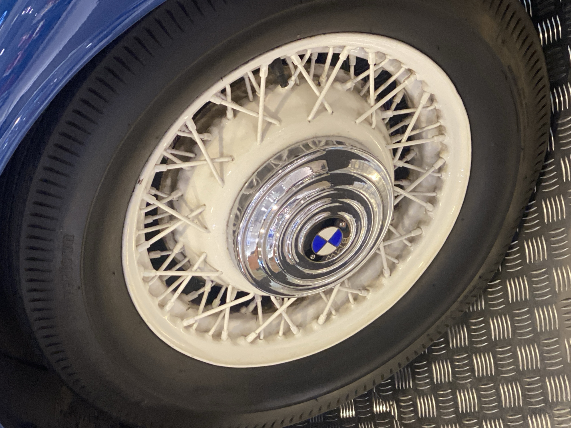 This is a close-up, eye-level shot of a spoked wheel from a vintage blue car, likely on display or in a showroom. The wheel is white with numerous thin spokes. In the center of the wheel is a chrome hubcap with a multi-layered, circular design. At the very center of the hubcap is a circular emblem with the BMW logo, featuring blue and white quadrants within a black circle, surrounded by the letters "BMW". The tire is black with a ribbed tread pattern. To the right of the wheel, a metallic floor with a diamond plate pattern is visible. The lighting is bright and artificial, suggesting an indoor setting. There is no visible text on the tire or the surrounding elements, except for the "BMW" lettering on the emblem. The image is sharp and focused on the wheel, with the car's blue bodywork visible in the upper left corner. The setting appears to be a professional display or exhibition, possibly related to automotive history or a car show, given the pristine condition of the vehicle and the specialized flooring. The location context is Łódź, Poland.