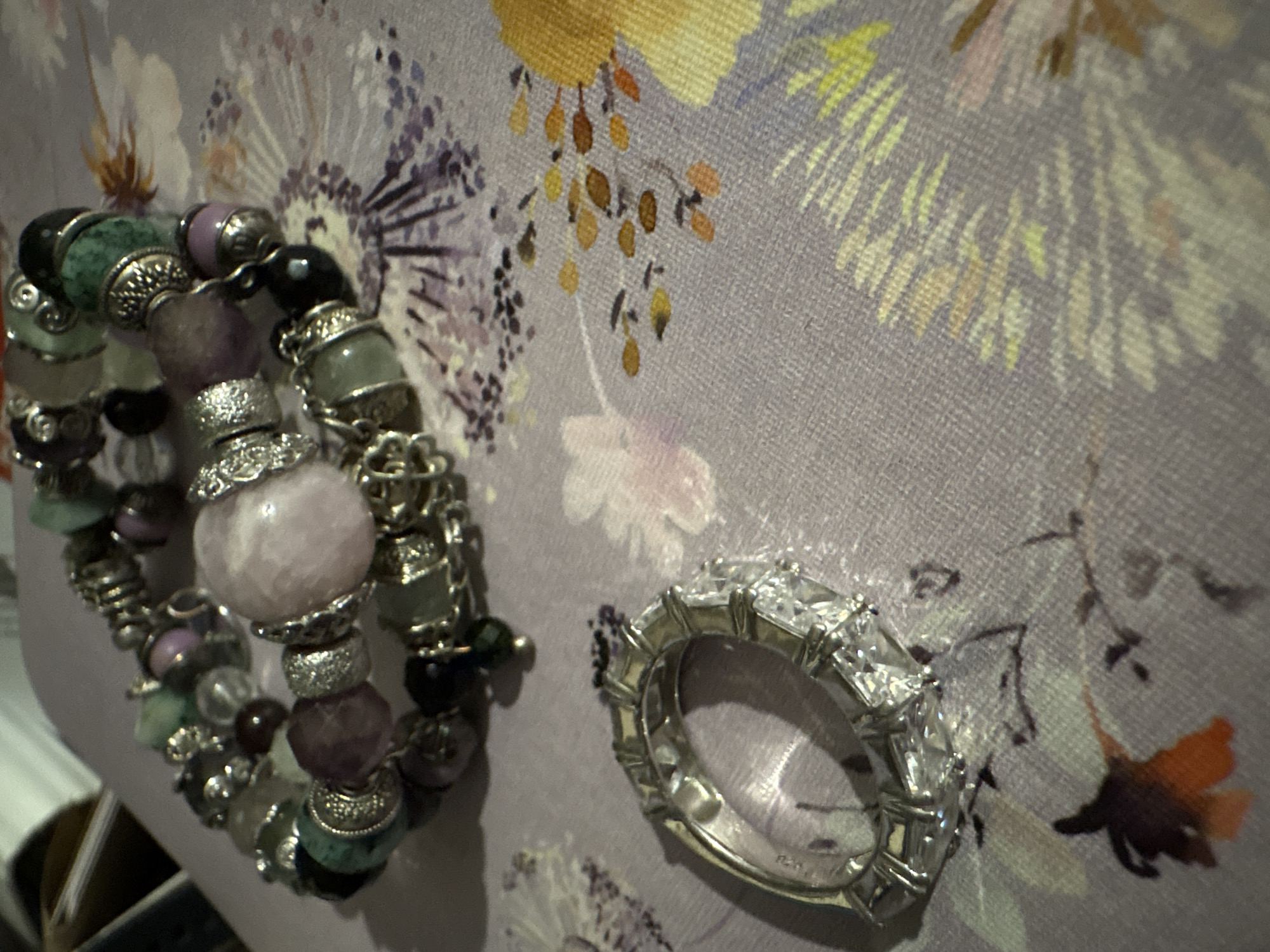 The image shows two pieces of jewelry resting on a patterned fabric. One is a multi-strand charm bracelet with various colored beads, metal spacers, and decorative elements. The other is a wide silver ring encrusted with rectangular clear stones. The fabric has a light purple background with a floral design that includes yellow, white, and purple flowers and brown leaves. The location and city of Izhevsk, Russia is not determinable from the context of this image.