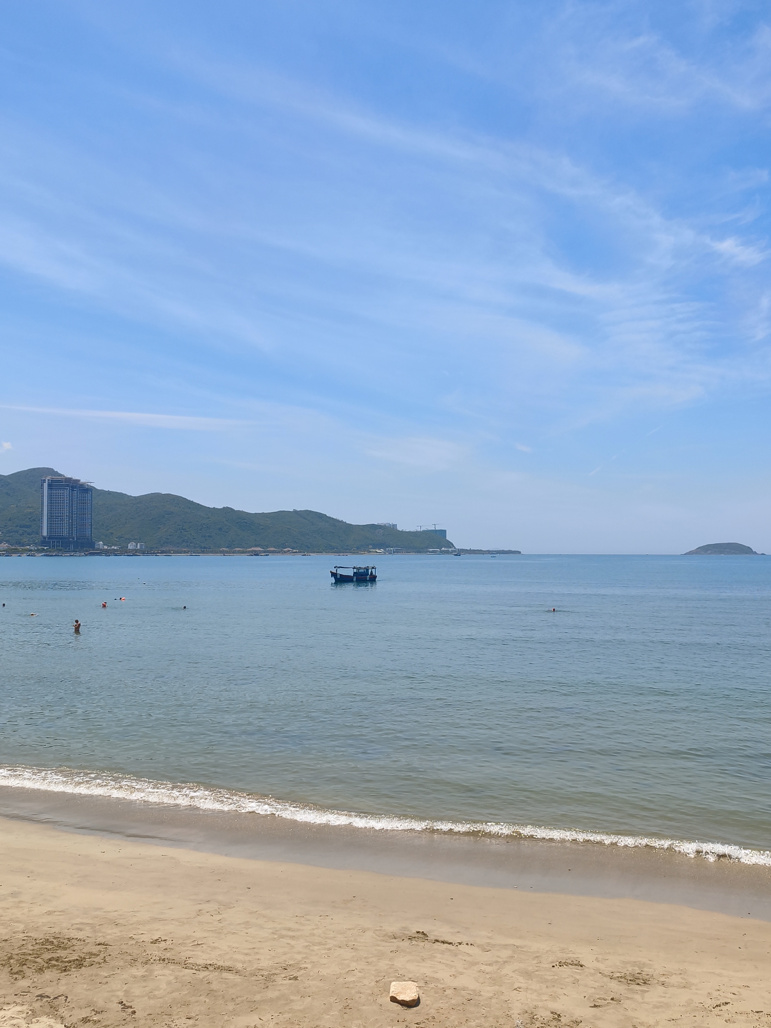 This is a medium shot, eye-level photograph taken from a sandy beach, looking out at the sea towards a distant shoreline with lush green hills. The sky is a clear, bright blue with thin, wispy white clouds scattered across it.
In the foreground, a tan-colored sandy beach stretches across the bottom of the frame. A thin line of foamy white waves gently laps at the shore.
The middle ground is occupied by the calm, blue sea. Several people are visible in the water, some swimming, some standing. A small, dark-colored boat with a canopy is anchored in the middle of the sea, with a few people on board. Further out, a lone person is visible swimming.
The background features a long stretch of coastline with rolling green hills. A tall, modern, multi-story building with a dark facade is prominently situated on the left side of the coastline, among other smaller structures and some construction cranes. The sea extends to the horizon, where another small, rounded island is visible on the far right.
The scene depicts a serene day at the beach in Nha Trang, Vietnam, likely a popular tourist spot given the modern development along the coast and the presence of swimmers and a boat. The overall atmosphere is peaceful and idyllic.