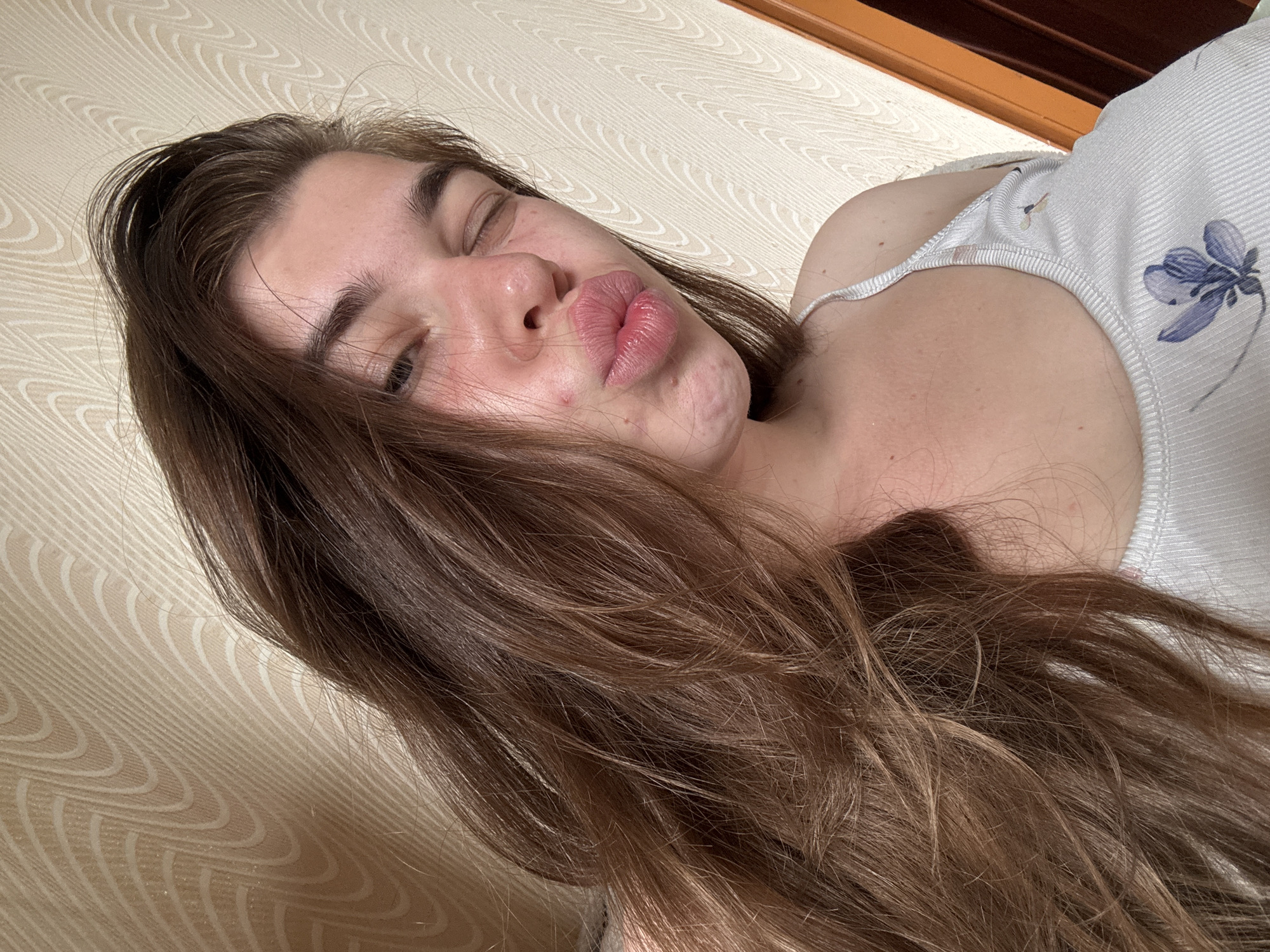 A young woman with long, brown, wavy hair is captured in a close-up, selfie-style shot. She is winking, with her lips pursed into a pout. Her face shows some minor blemishes on her chin and cheek. She is wearing a white ribbed tank top with a subtle floral pattern. The background appears to be a wall with a textured, light-colored wallpaper featuring an abstract, wavy pattern. To the right, a portion of a wooden doorframe is visible. The lighting suggests an indoor setting, possibly a bedroom, with natural light filtering in. There is no specific indication of the city of Мещёры, Russia, in the visual elements of the image.
