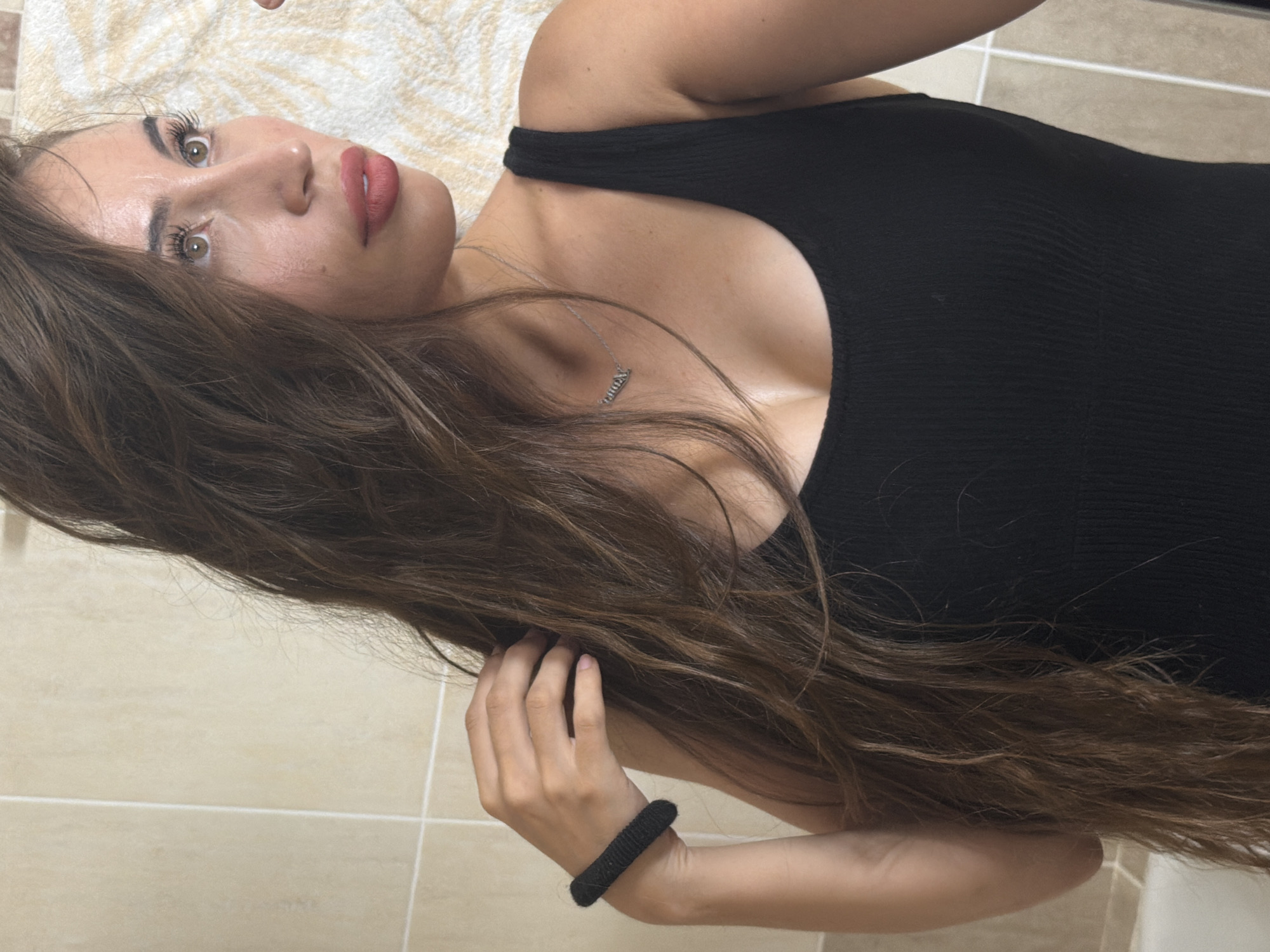 A medium shot photo shows a young woman with long brown hair and a black tank top. Her hair is long and wavy and cascades down her right shoulder. She is wearing a black beaded bracelet on her left wrist and a silver necklace with a pendant that spells out a name. She has on a light pink lipstick and her eyes are open and looking to the right. The background is a wall with beige tiles and a towel with a white and tan pattern. The photo appears to be taken in a bathroom. The woman is in the city of Tersefanou, Cyprus.