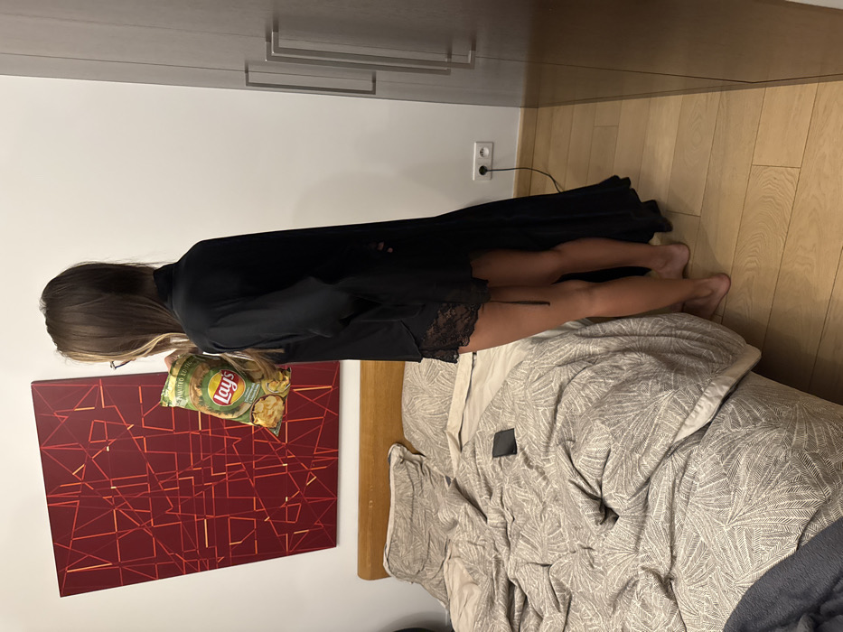 A woman stands at the foot of a bed with a bag of Lays chips in hand. She wears a black robe, revealing lace lingerie. The bed is made with a patterned grey and white comforter and headboard visible. A red abstract art piece hangs on the wall near the bed. Wood flooring extends into the corner of the room. A white dresser stands against the wall, next to an electrical outlet.